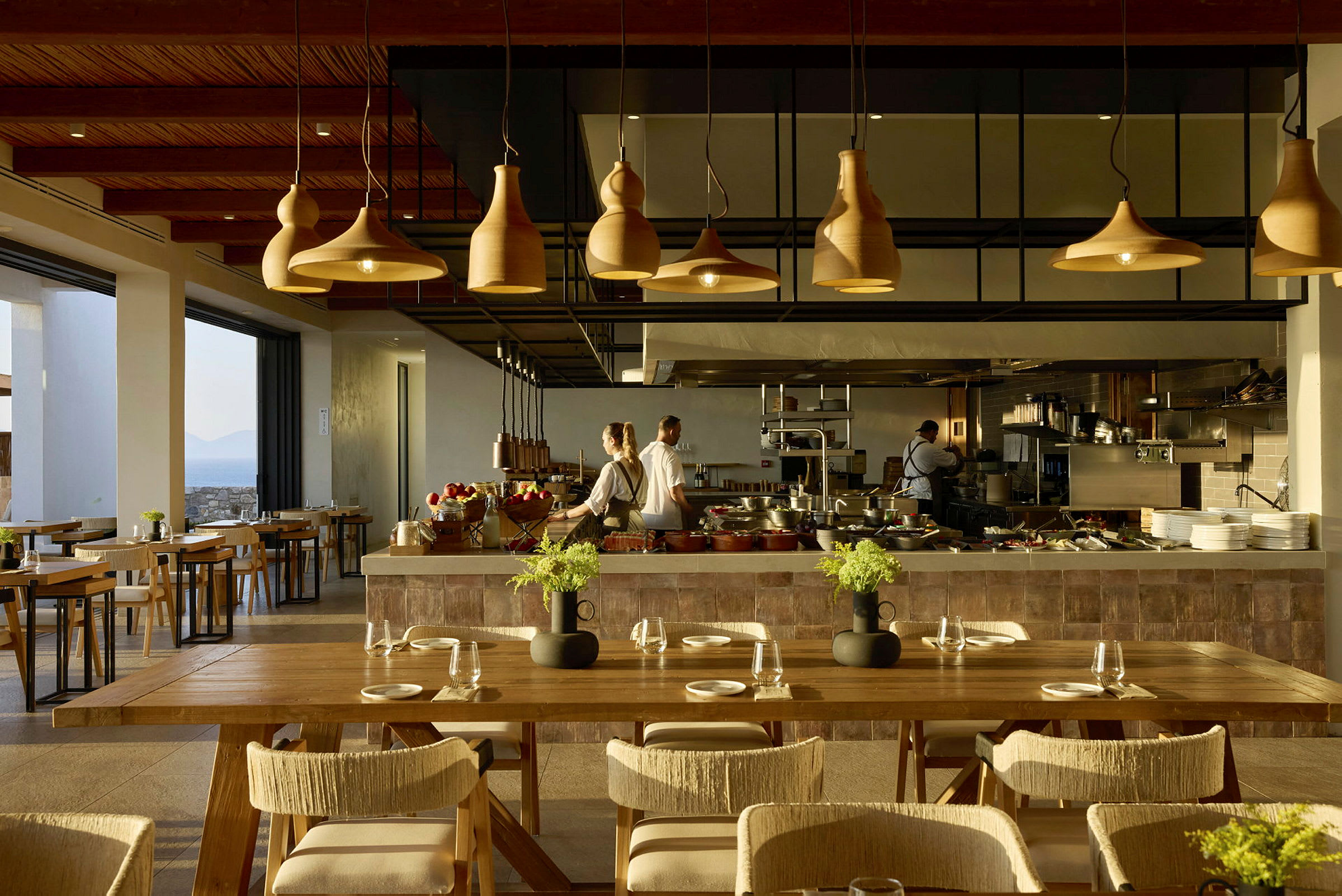 Et moderne restaurantmiljø med elegante træ- og lysdesigndetaljer, hvor kokke arbejder bag en serveringsdisk. (AI-genereret tekst)