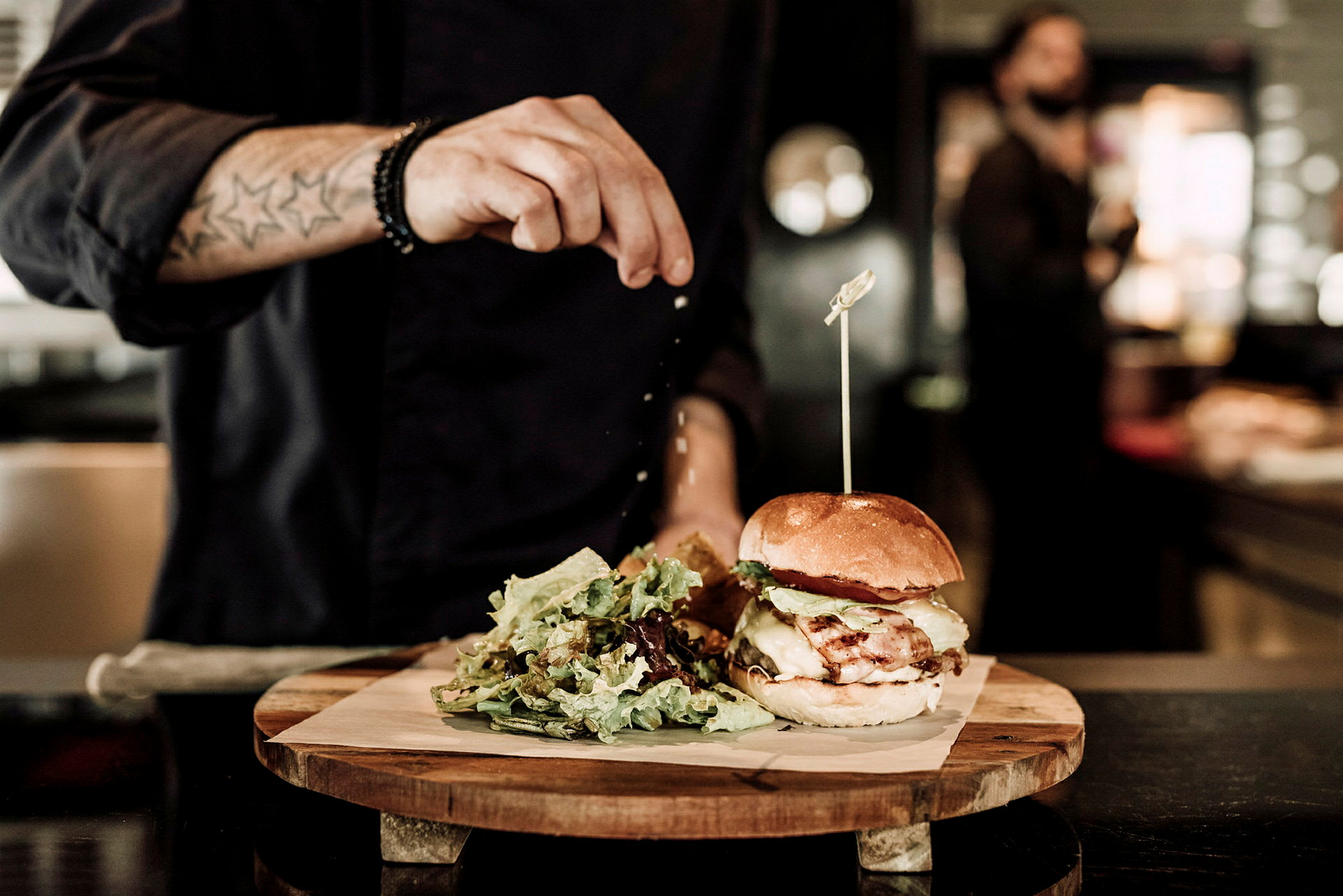 En person krydrer en burger, som er placeret på en træplade sammen med en portion salat. (AI-genereret tekst)