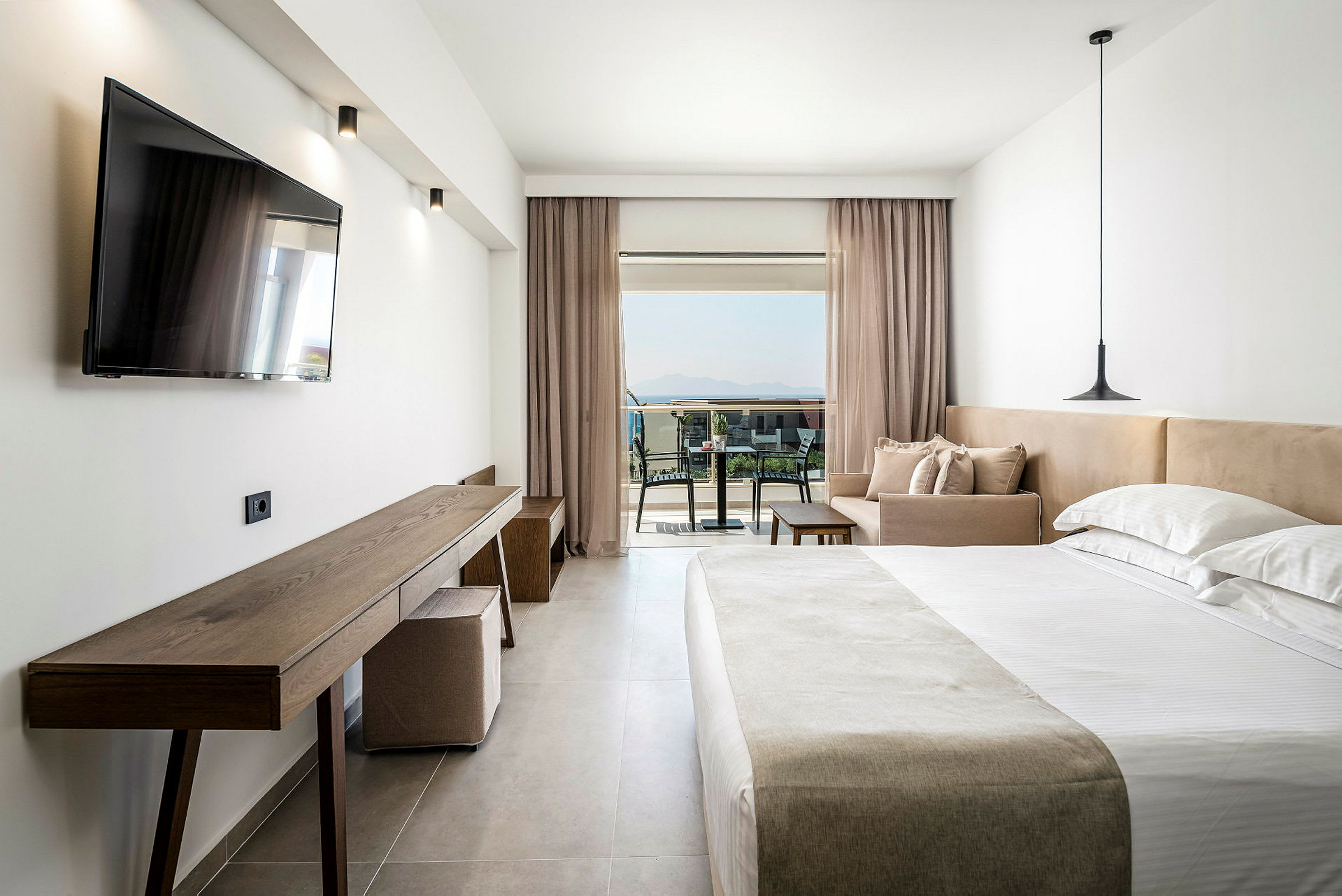 Interiør af et hotelværelse med dobbeltseng, sofa og skrivebord. Bag glasdøre ses en møbleret balkon med udsigt over omgivelserne. (AI-genereret tekst)