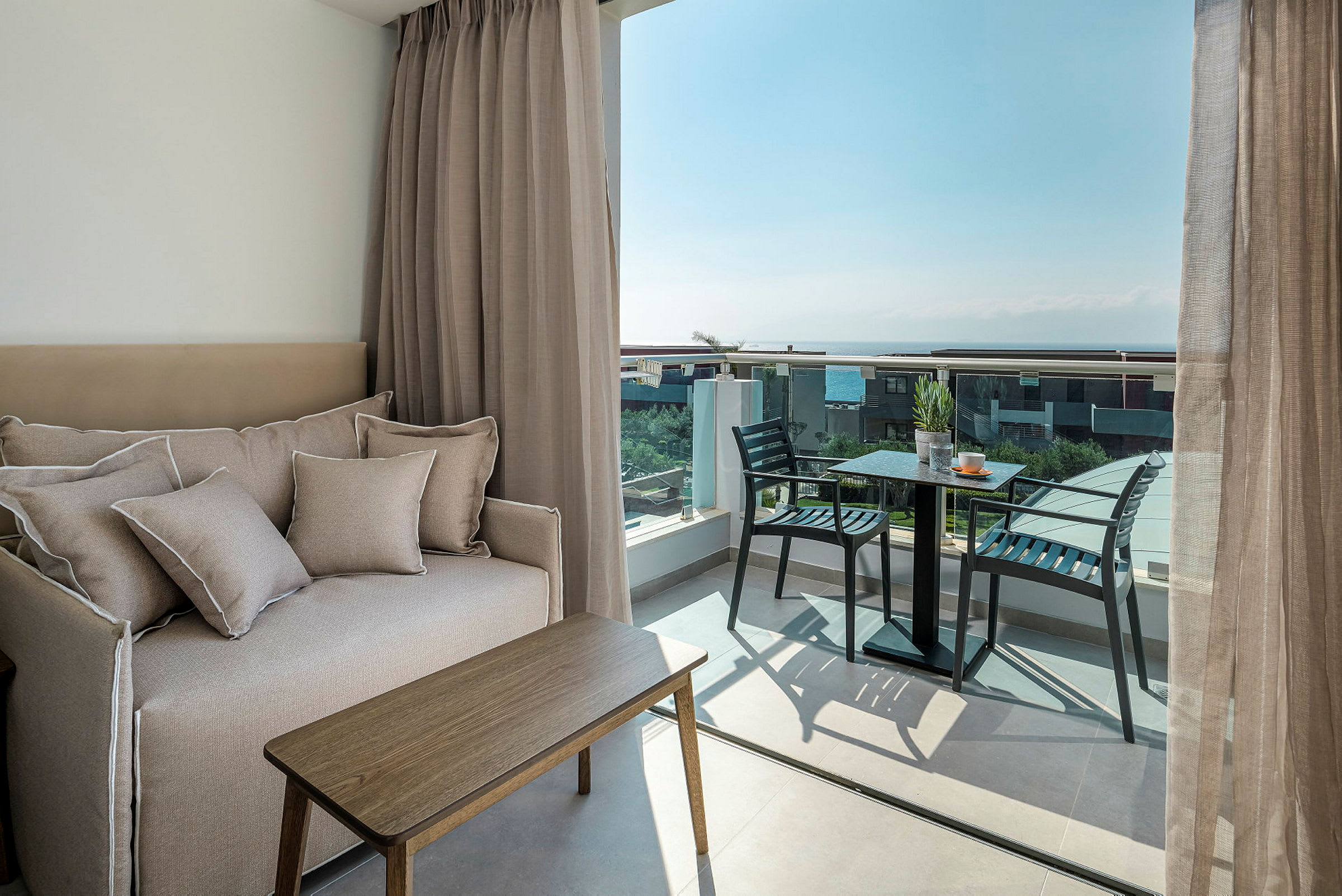 Billede af et hotelværelse med en sofa, et lille bord og en altan med stole og bord samt udsigt over havet. (AI-genereret tekst)