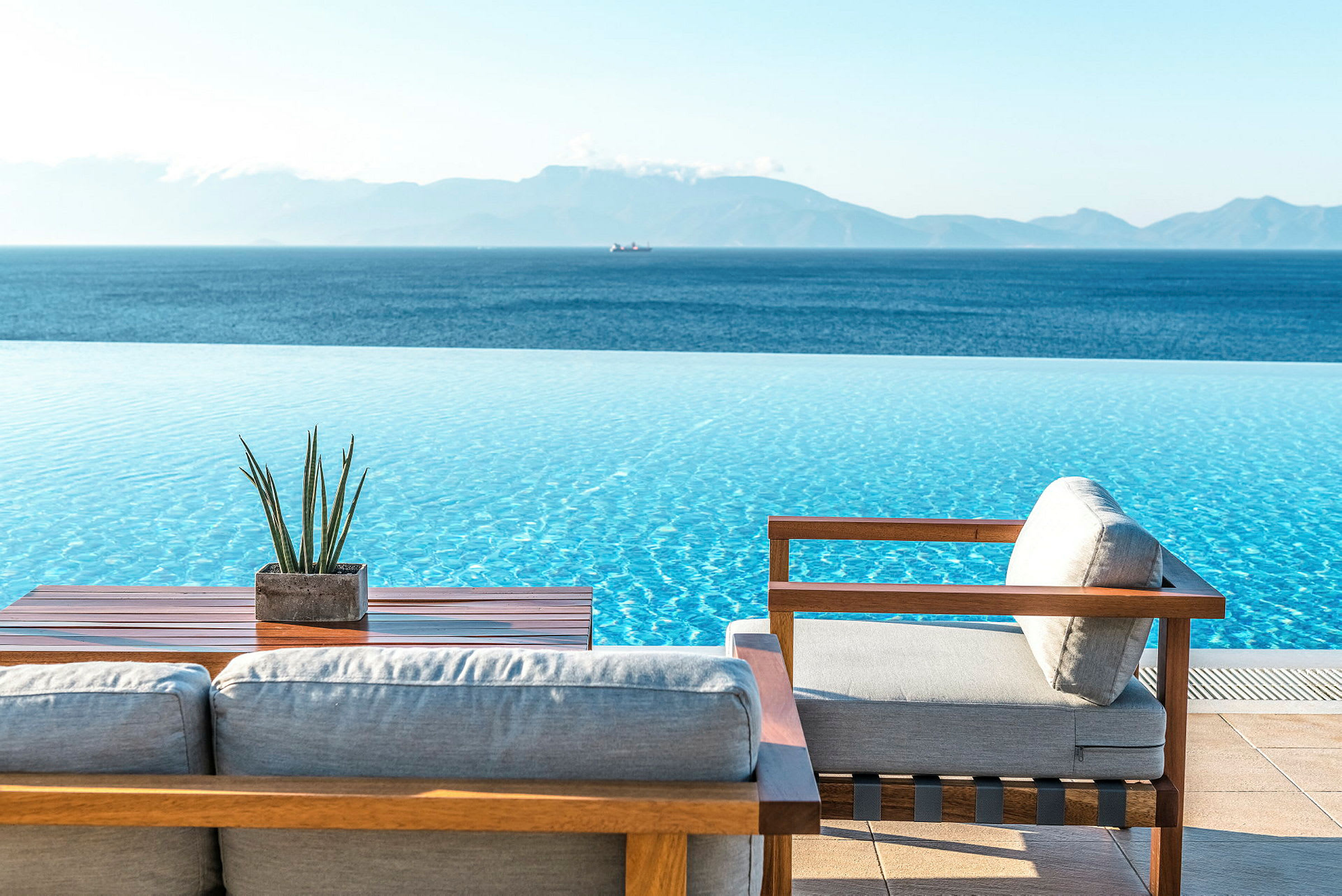 En infinity-pool med udsigt over en blå hav og fjerne bjerge, med træmøbler og en lille plante på et bord i forgrunden. (AI-genereret tekst)