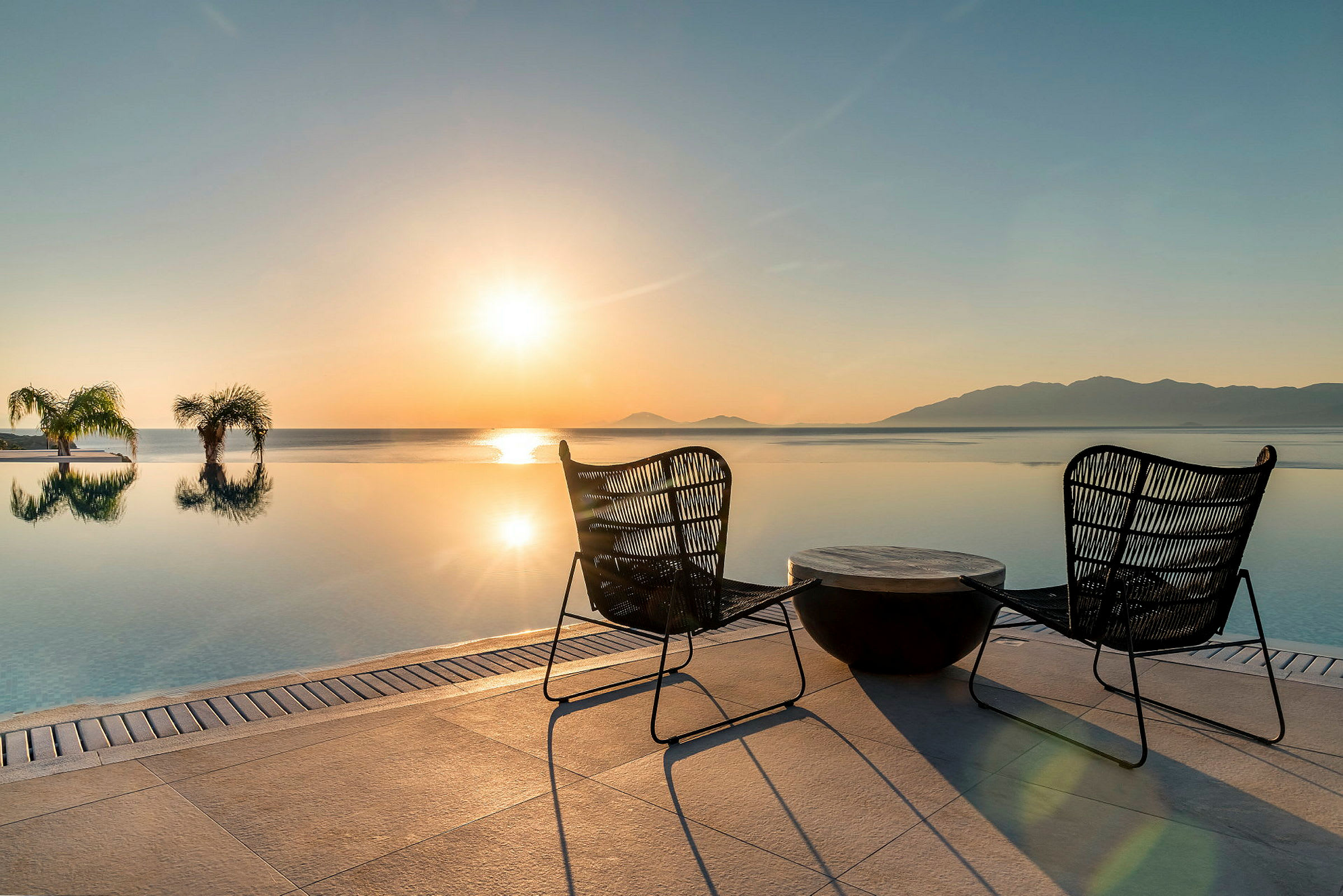 To stole og et bord ved en infinitypool med udsigt over havet i solnedgang. (AI-genereret tekst)