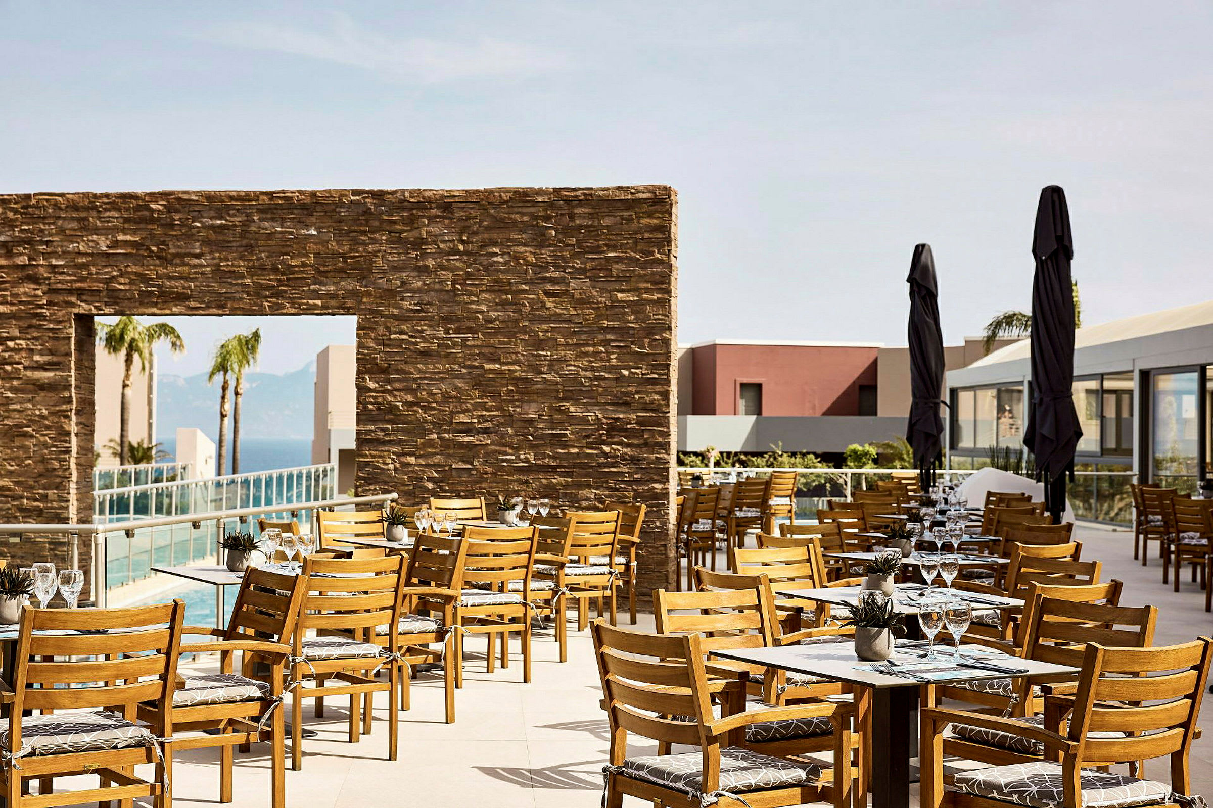 En udendørs restaurant med træstole og borde, omgivet af en moderne bygning og en pool. (AI-genereret tekst)