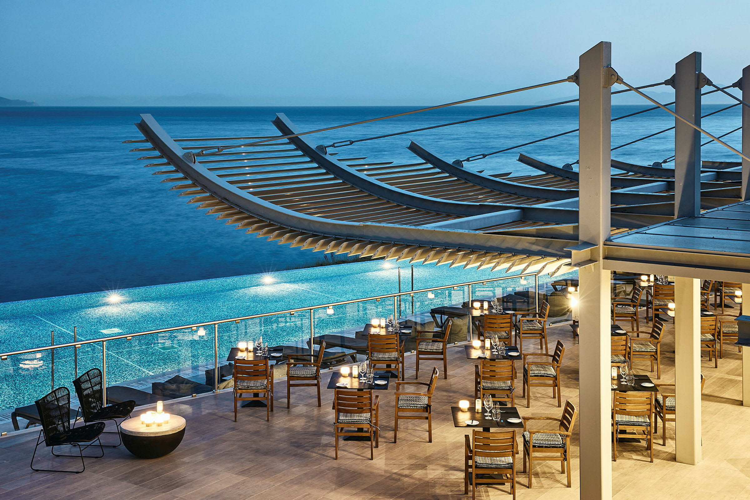 Restaurant med træstole og borde ved en pool, havet i baggrunden. (AI-genereret tekst)