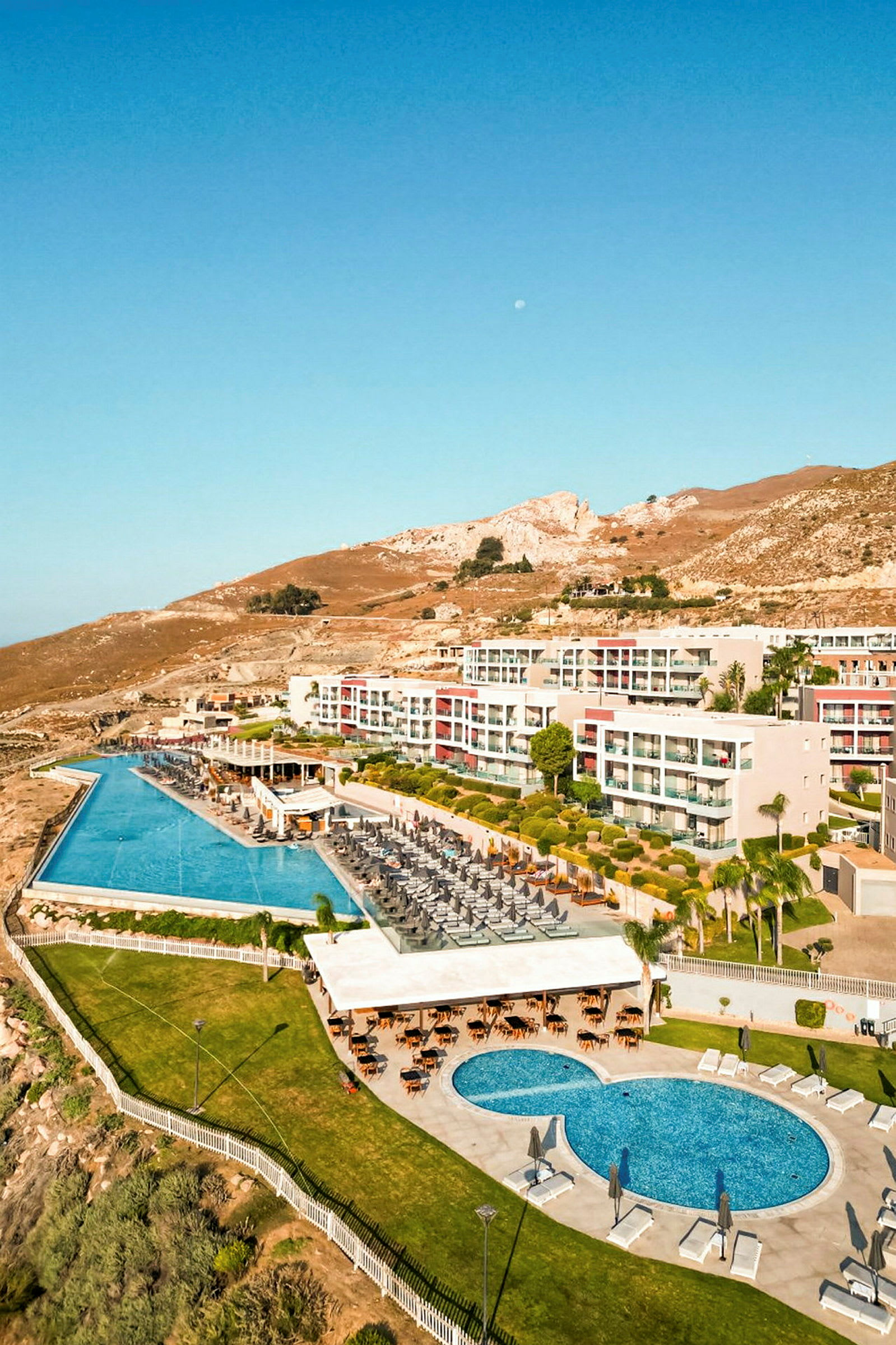 Billede af et hotelområde med pools, liggestole og grønt, beliggende ved et bakket landskab. (AI-genereret tekst)