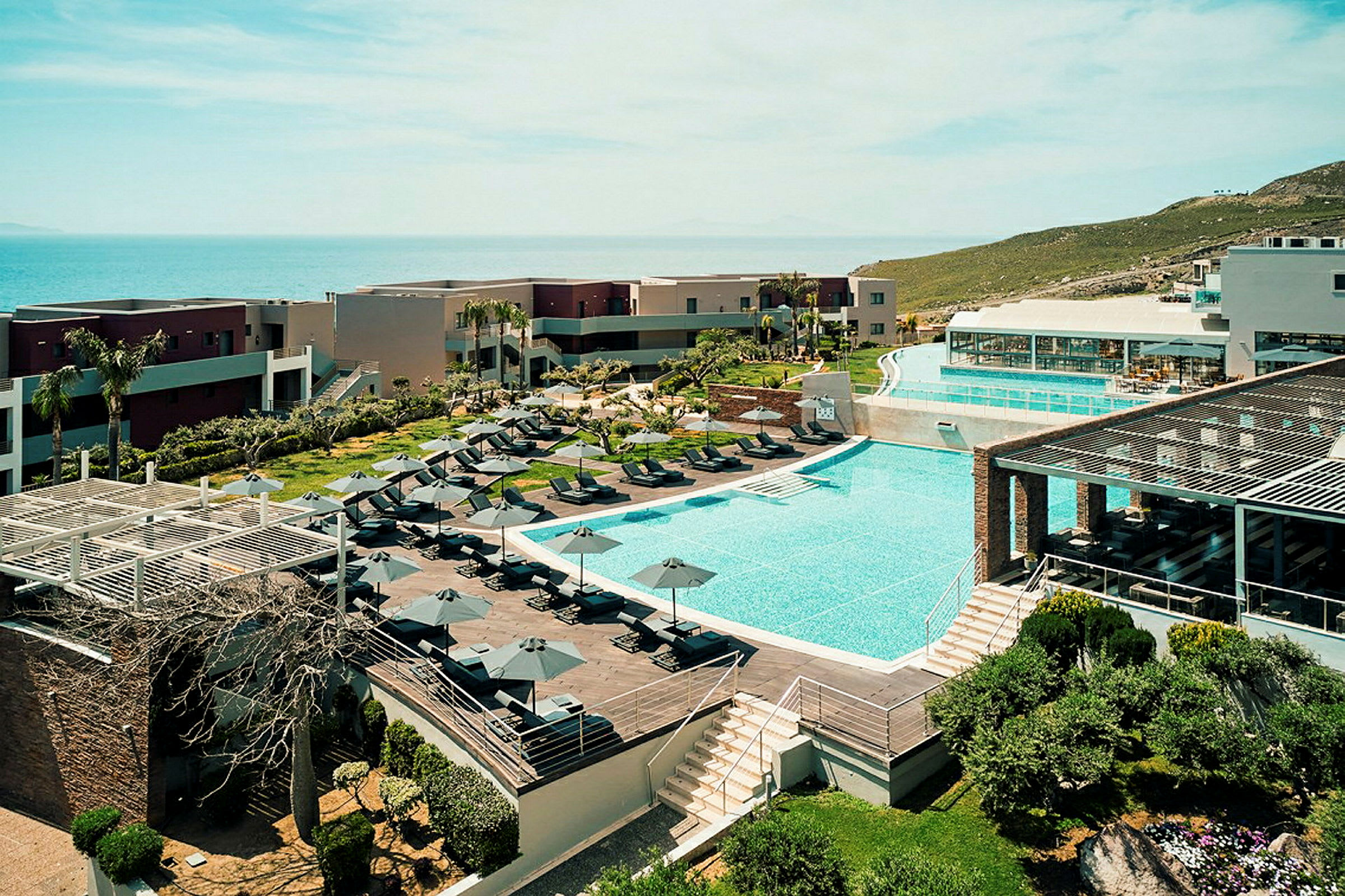 Hotelanlæg med pool, liggestole og omgivende bygninger ved havet. (AI-genereret tekst)