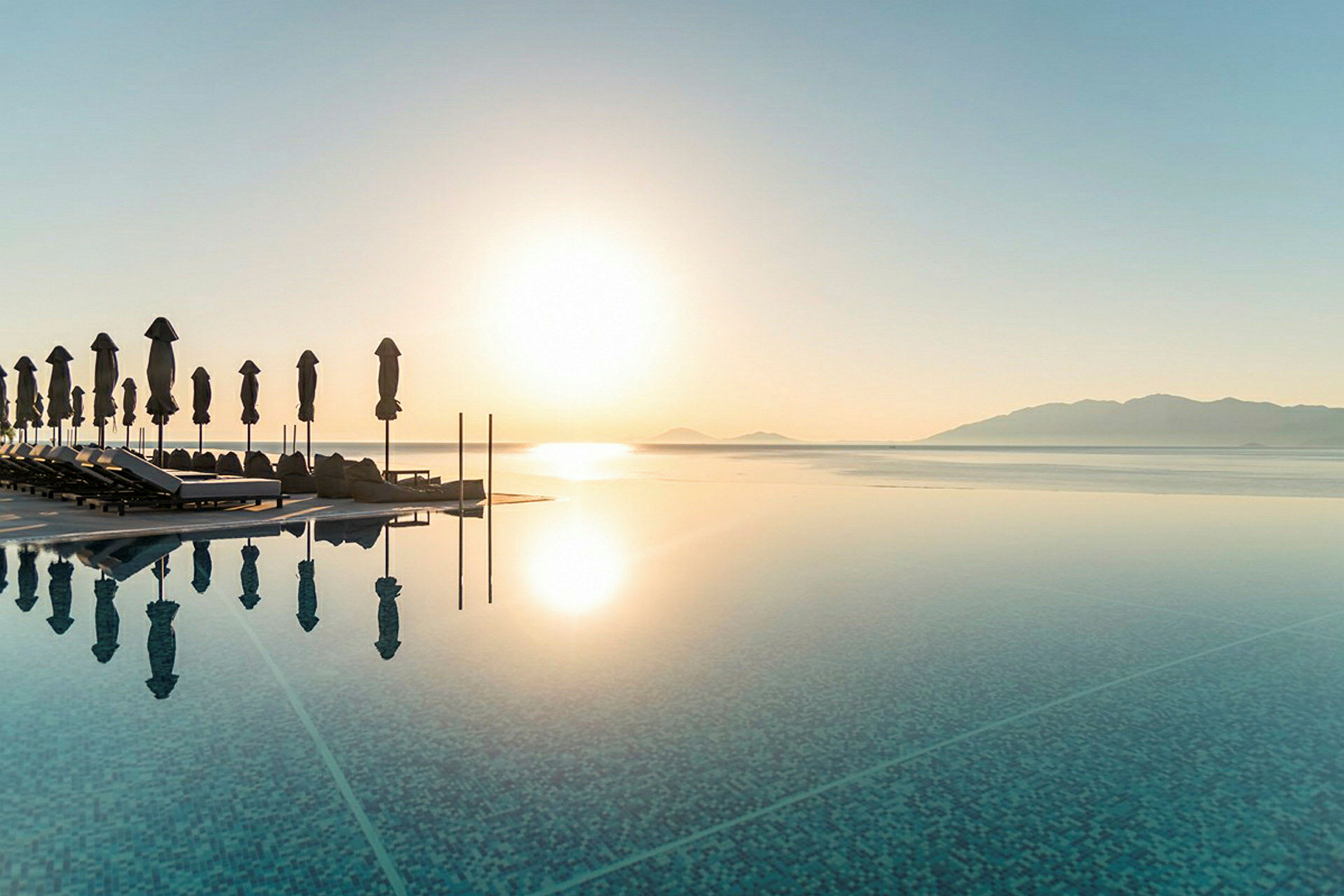 Strand med liggestole og parasoller ved en infinitypool med udsigt over havet og bjerge i solnedgangen. (AI-genereret tekst)