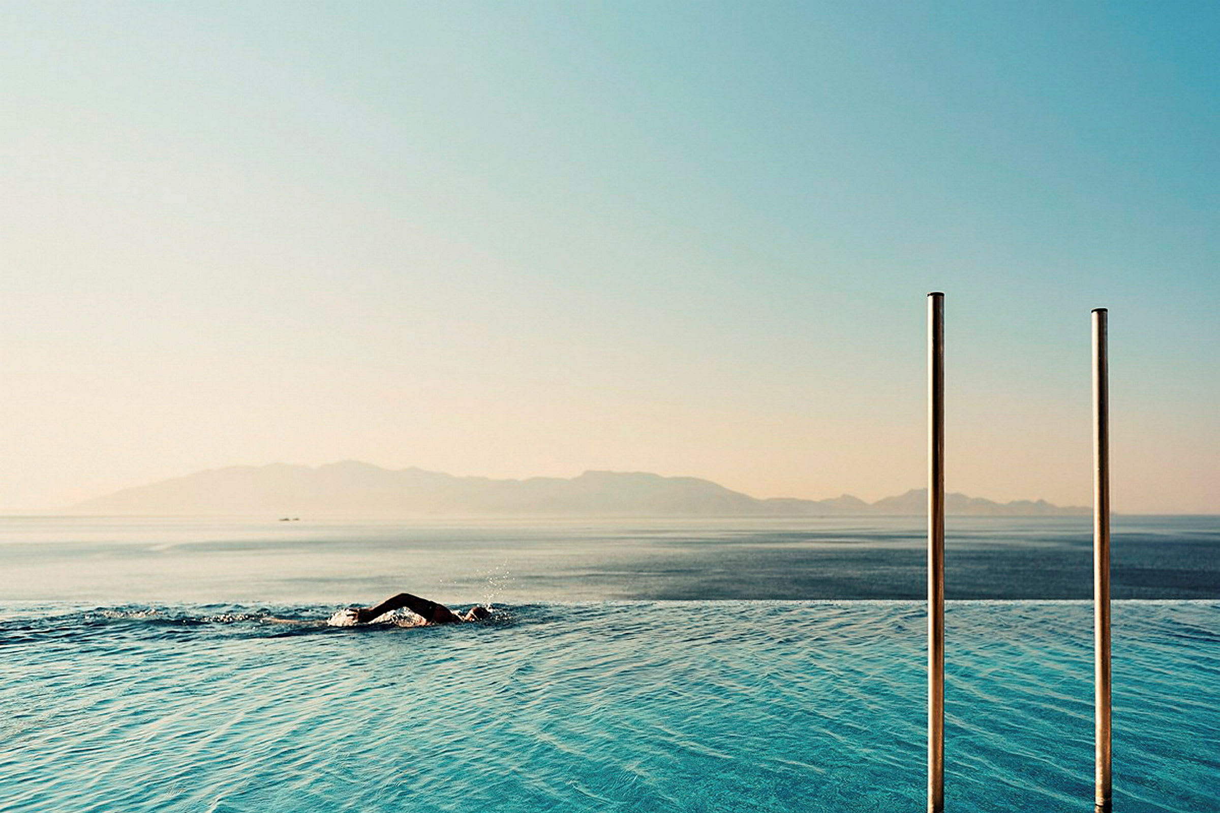 En person svømmer i en infinitypool med havet og bjerge i baggrunden. (AI-genereret tekst)