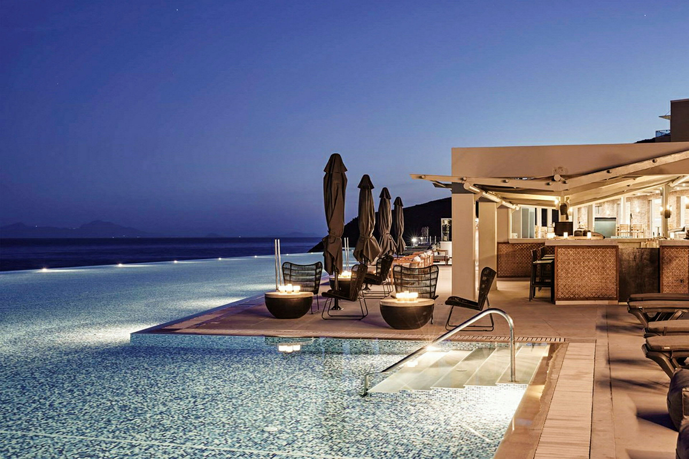 En pool ved siden af en restaurant med siddepladser og en fantastisk udsigt under en blå nattehimmel. (AI-genereret tekst)