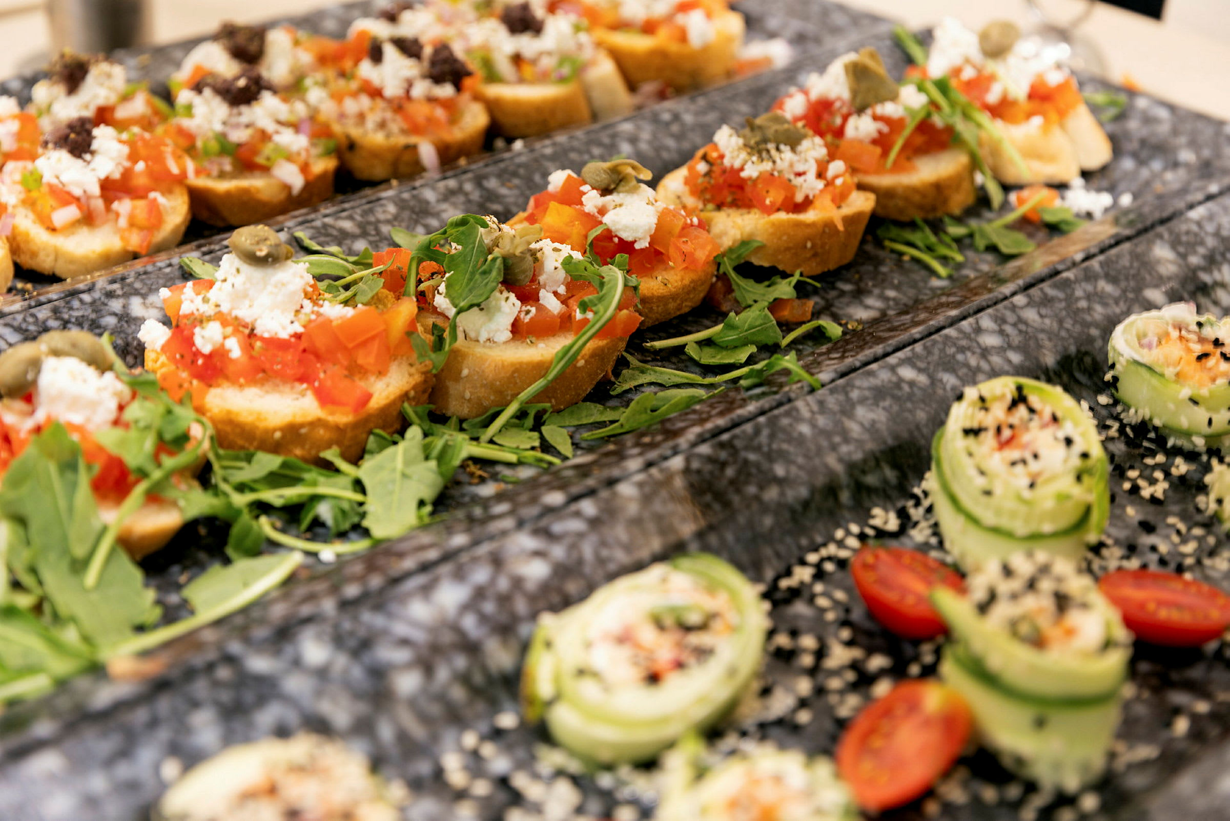 Anretning af bruschetta med tomat, fetaost og rucola samt små agurkesnitter med sesamfrø og cherrytomater. (AI-genereret tekst)