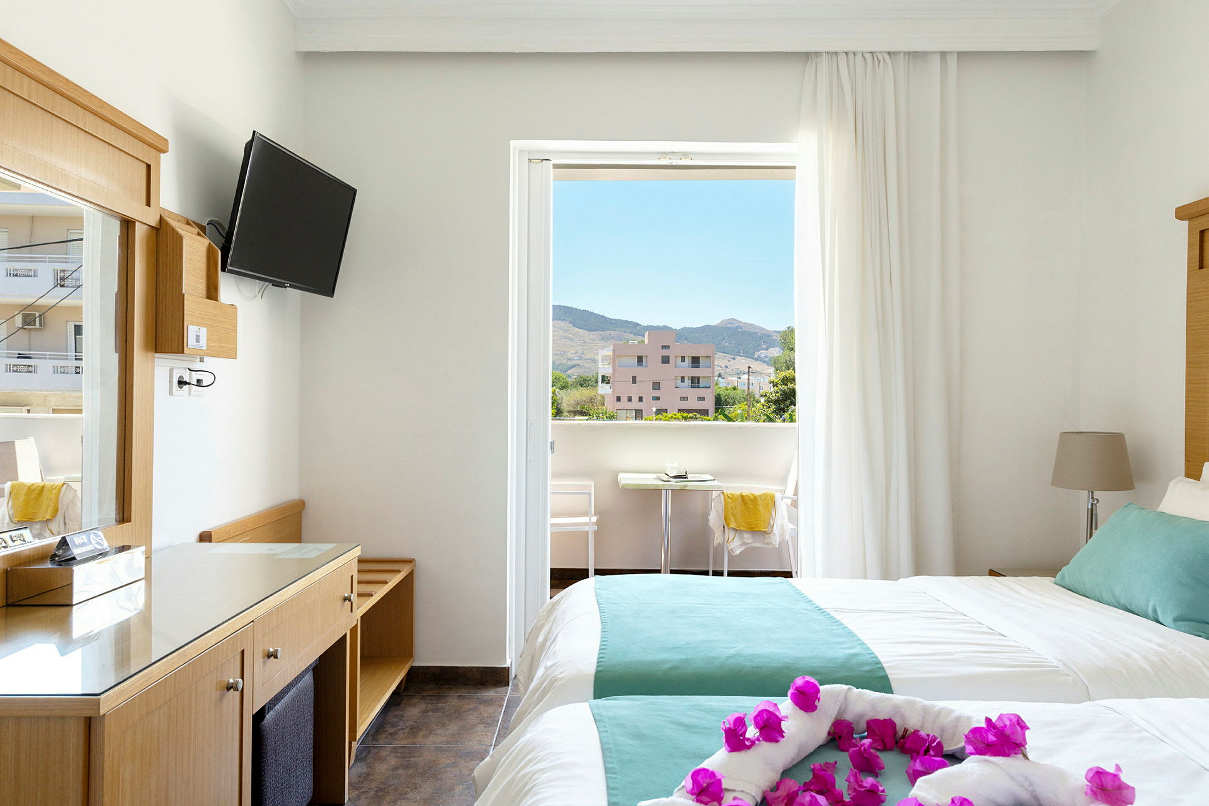 Hotelværelse med to senge og balkon. Udsigt over omkringliggende bygninger og bjerge. (AI-genereret tekst)