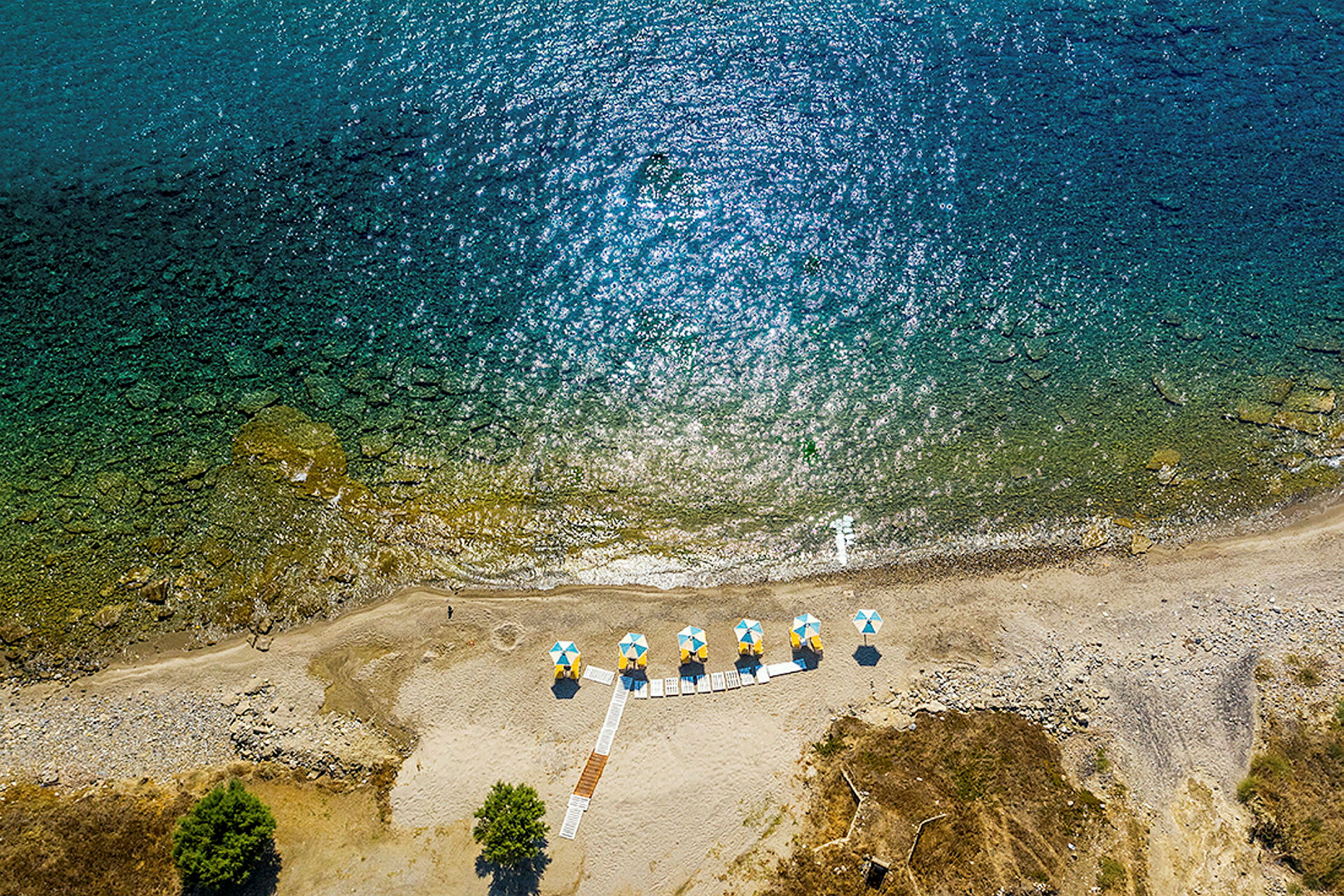 Luftfoto af en strand med parasoller i gule og blå farver samt gangstier. (AI-genereret tekst)