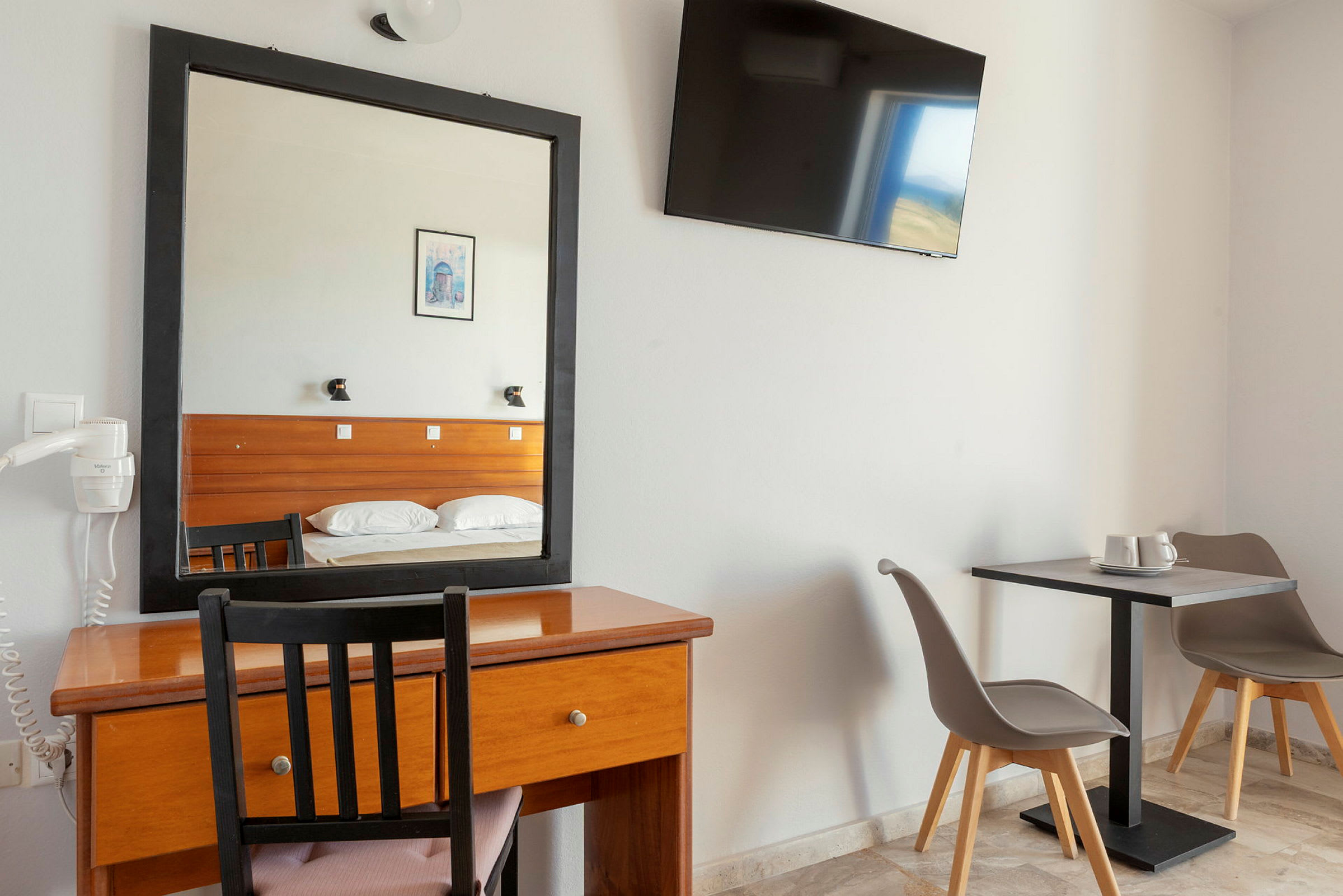 Hotelværelse med skrivebord, spejl og TV på væggen. Til højre to stole ved et bord. (AI-genereret tekst)