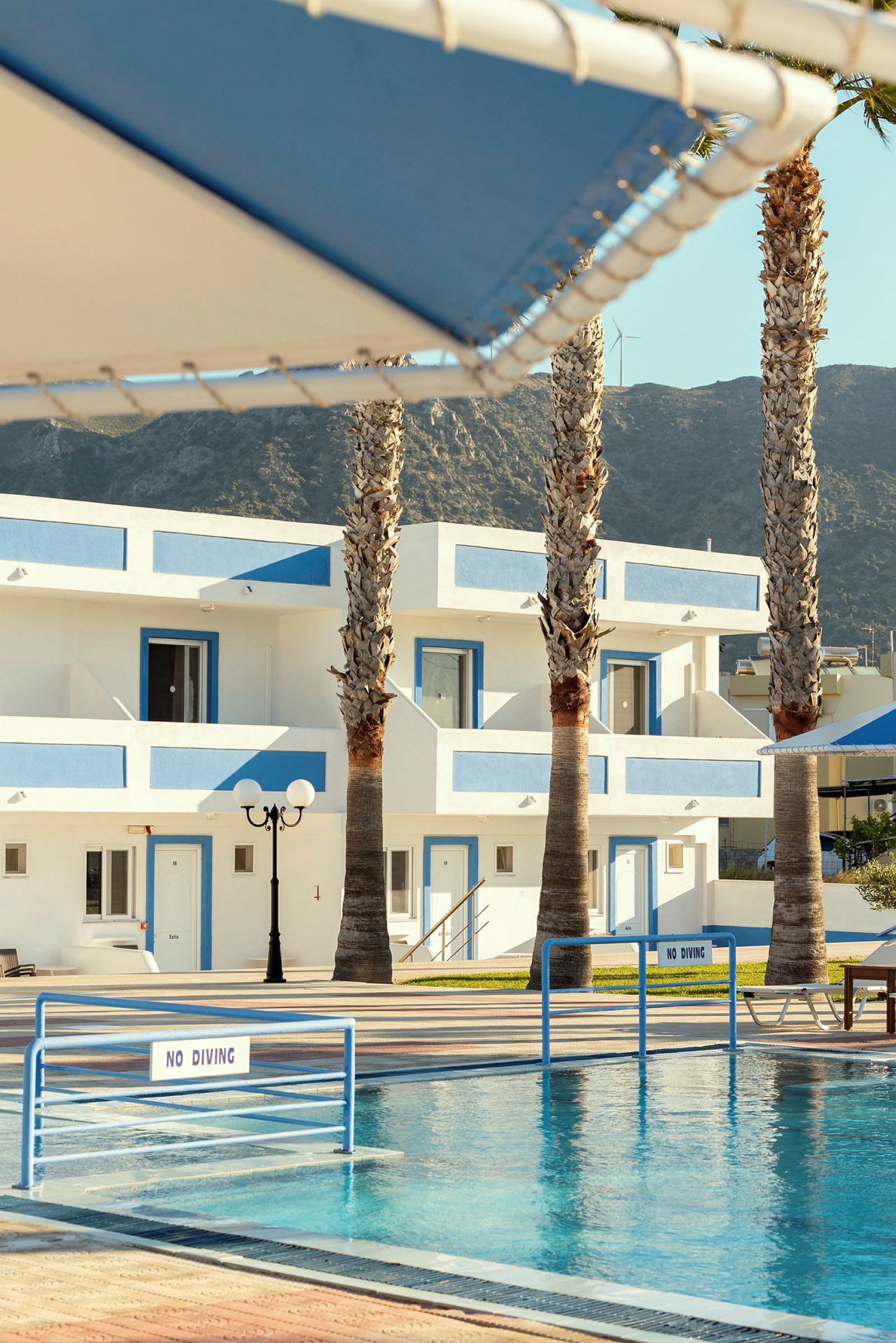 Hotelområde med hvid bygning og blå detaljer, pool og palmer. (AI-genereret tekst)