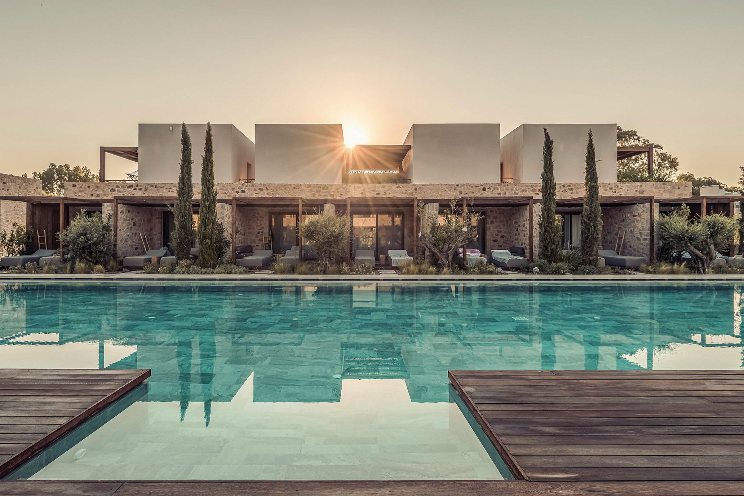 Billede af en moderne villa med en pool foran, omgivet af vegetation og træer, med solens stråler synlige bag bygningen. (AI-genereret tekst)
