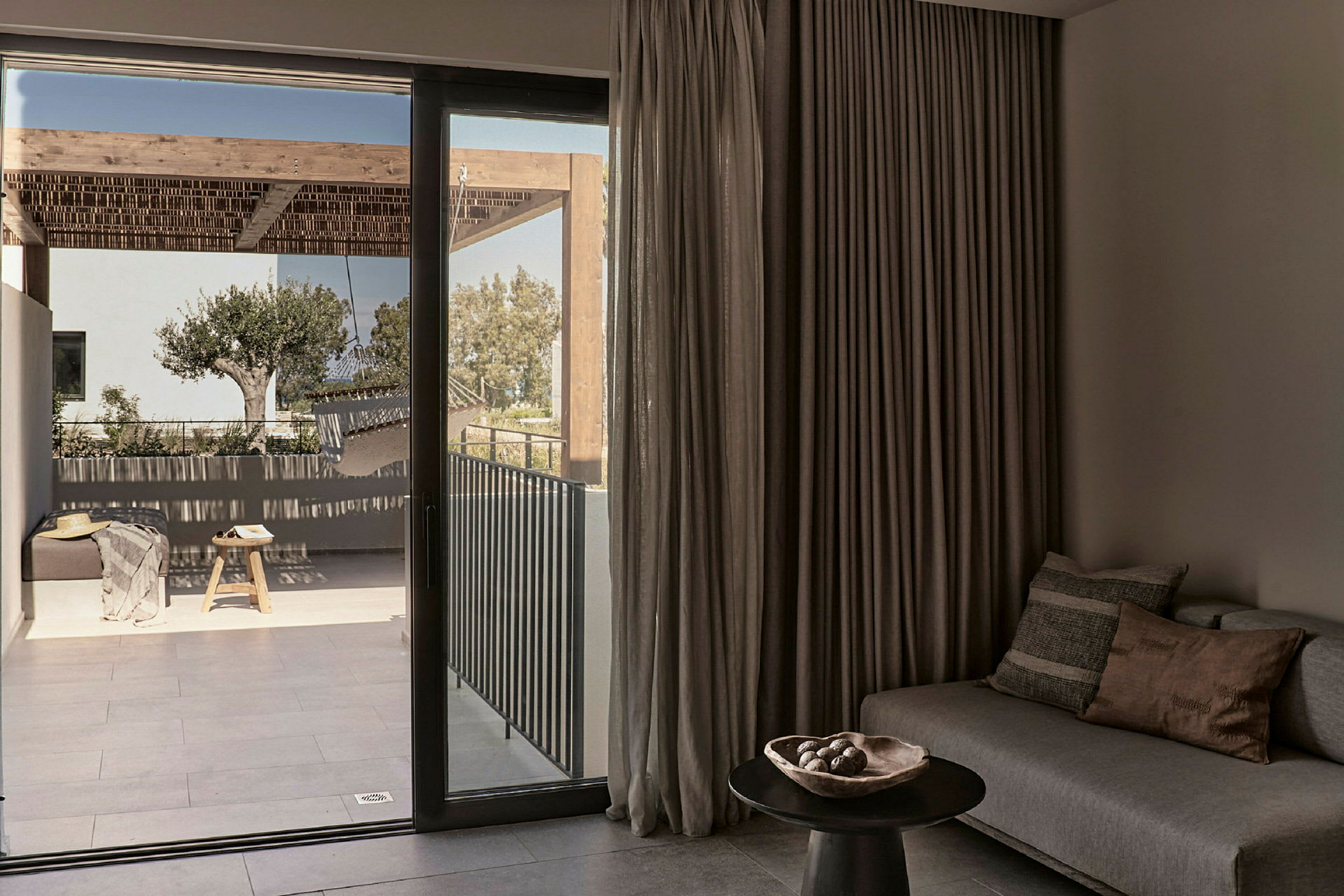 Inde i et rum med en sofa og et bord, set mod en terrasse med træmøbler og et træ. (AI-genereret tekst)