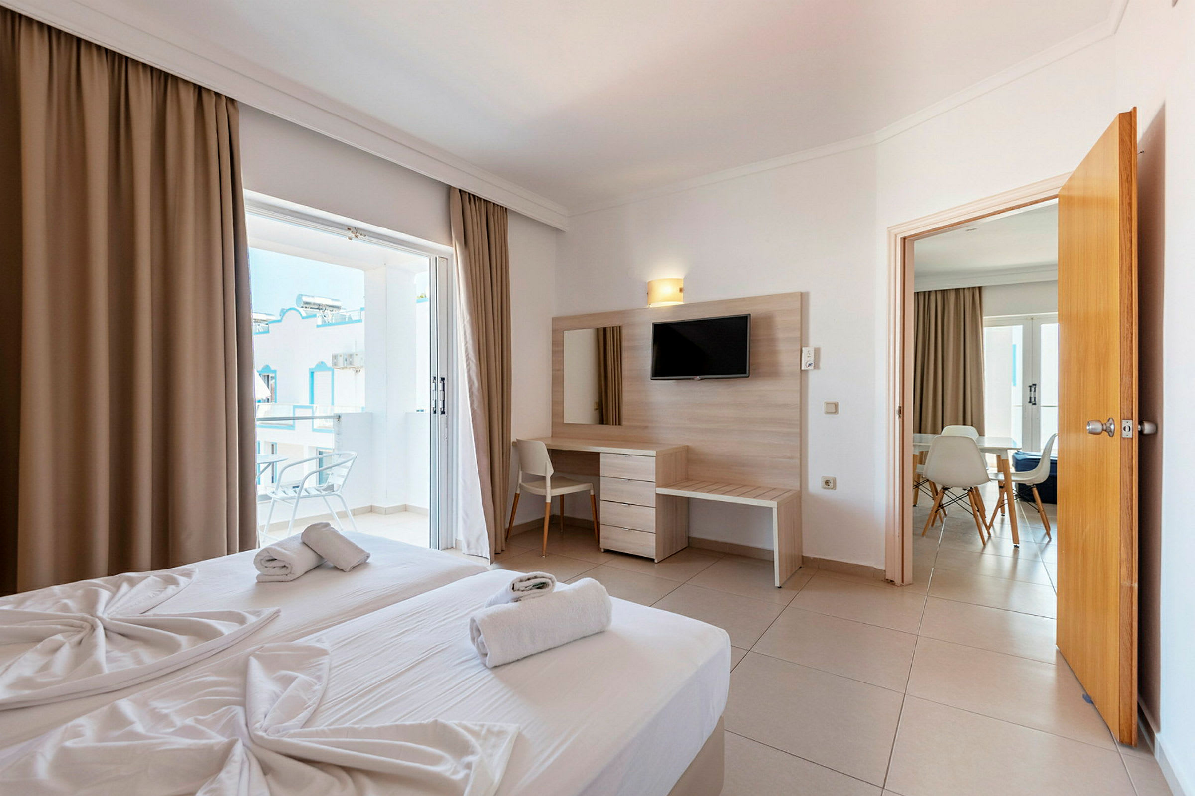 Moderne hotelværelse med to senge, et skrivebord med spejl og TV. Altan med udsigt til en hvid bygning. (AI-genereret tekst)