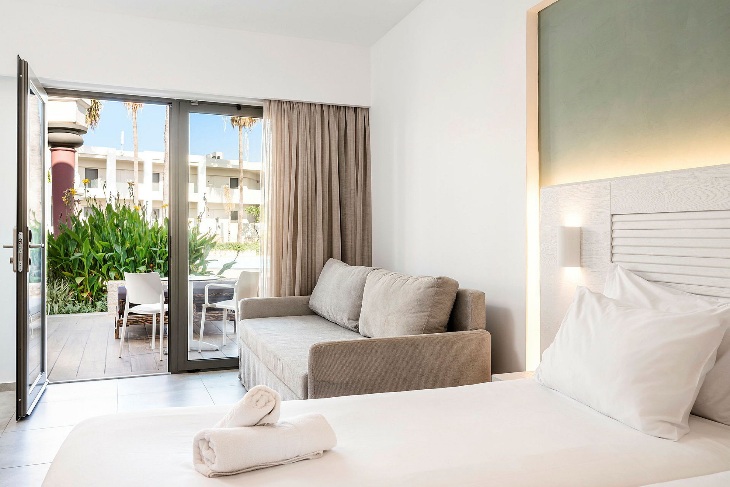 Et moderne hotelværelse med en dobbeltseng, en grå sofa og en åben glasdør, der fører til en terrasse med stole og grønt. (AI-genereret tekst)
