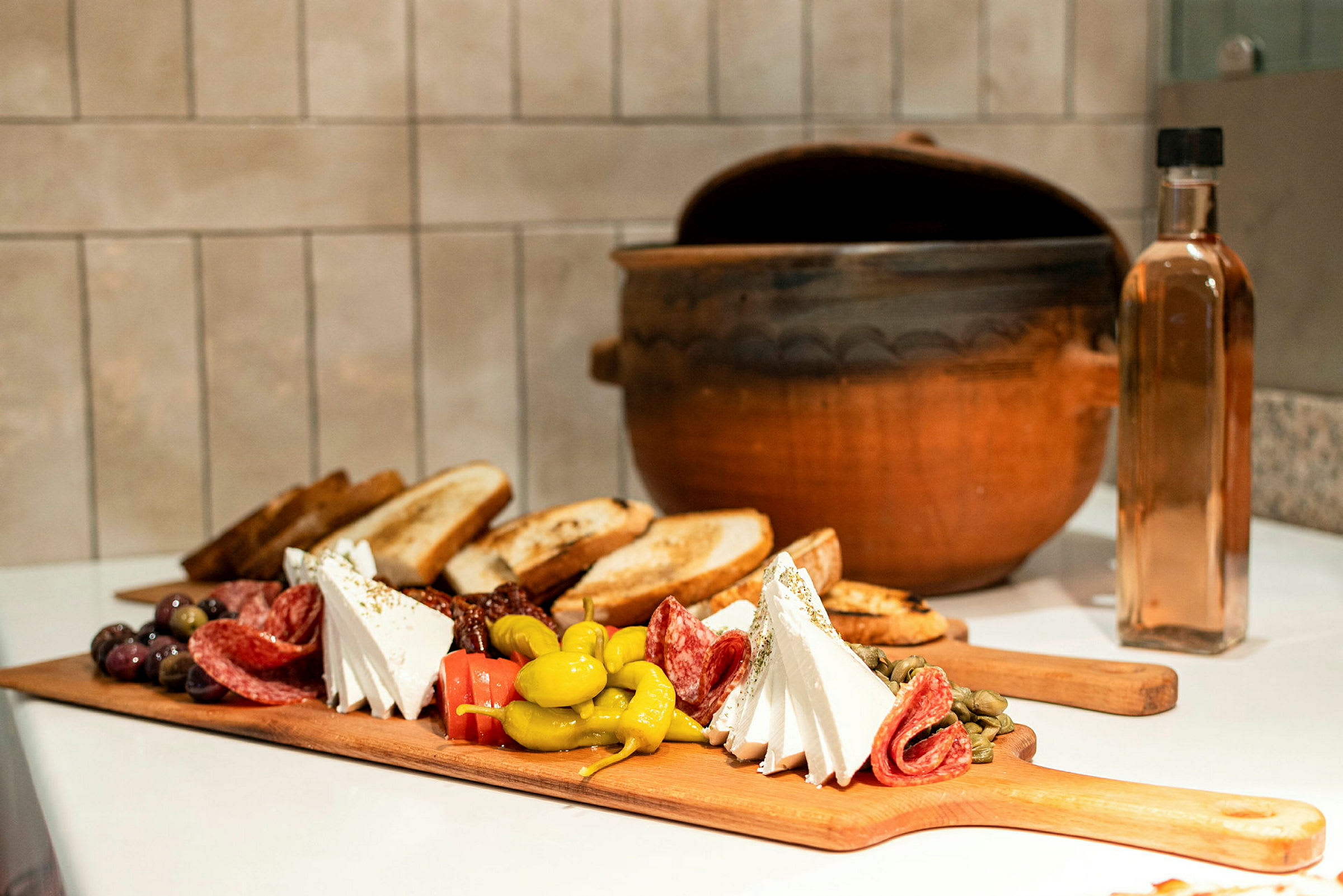 Et skærebræt med forskellige antipasti som ost, salami, oliven og brød, samt en lergryde og en glasflaske i baggrunden. (AI-genereret tekst)