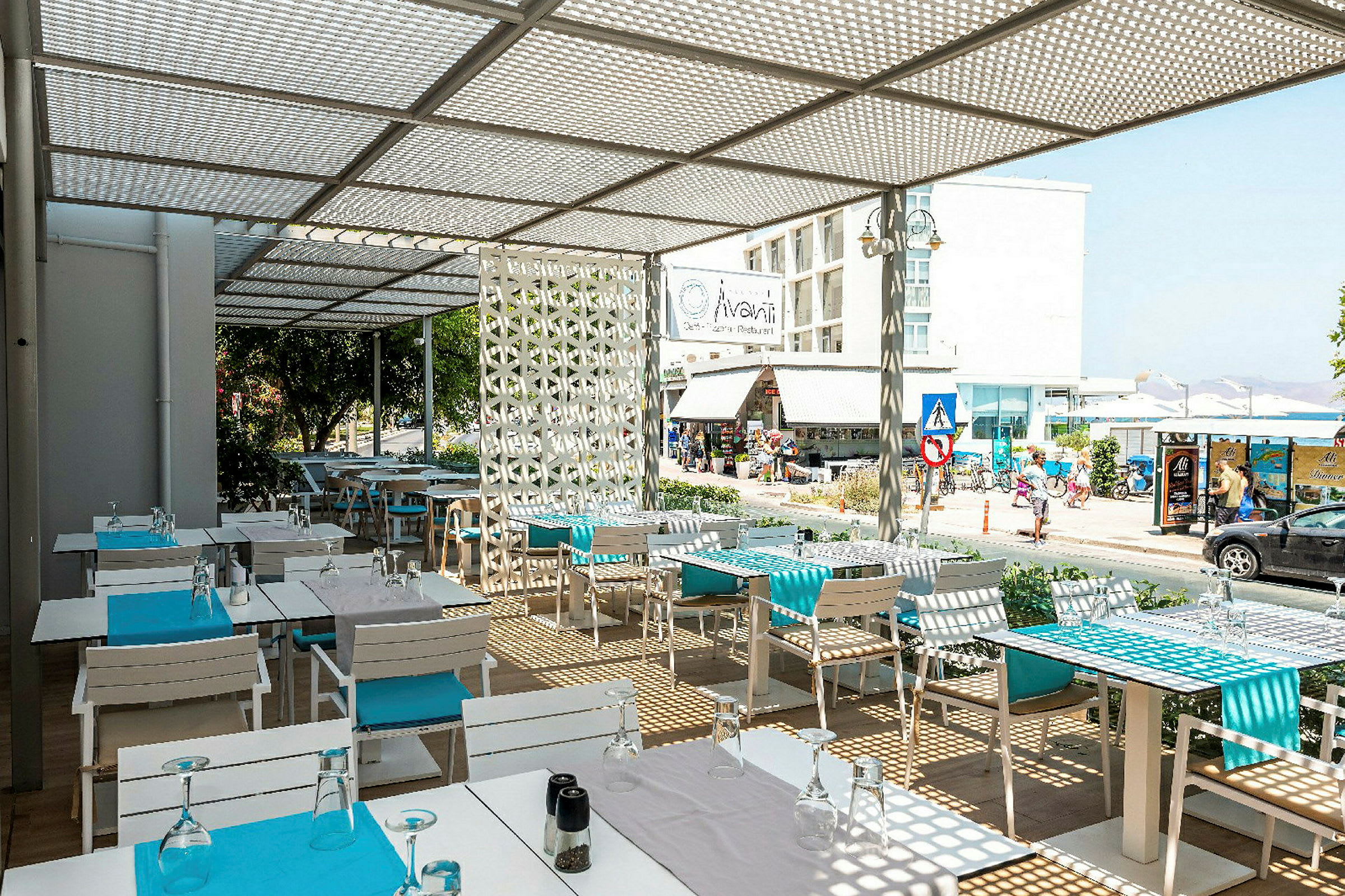 En udendørs restaurant med hvide borde og blå detaljer, med udsigt til en gade og bygninger. (AI-genereret tekst)