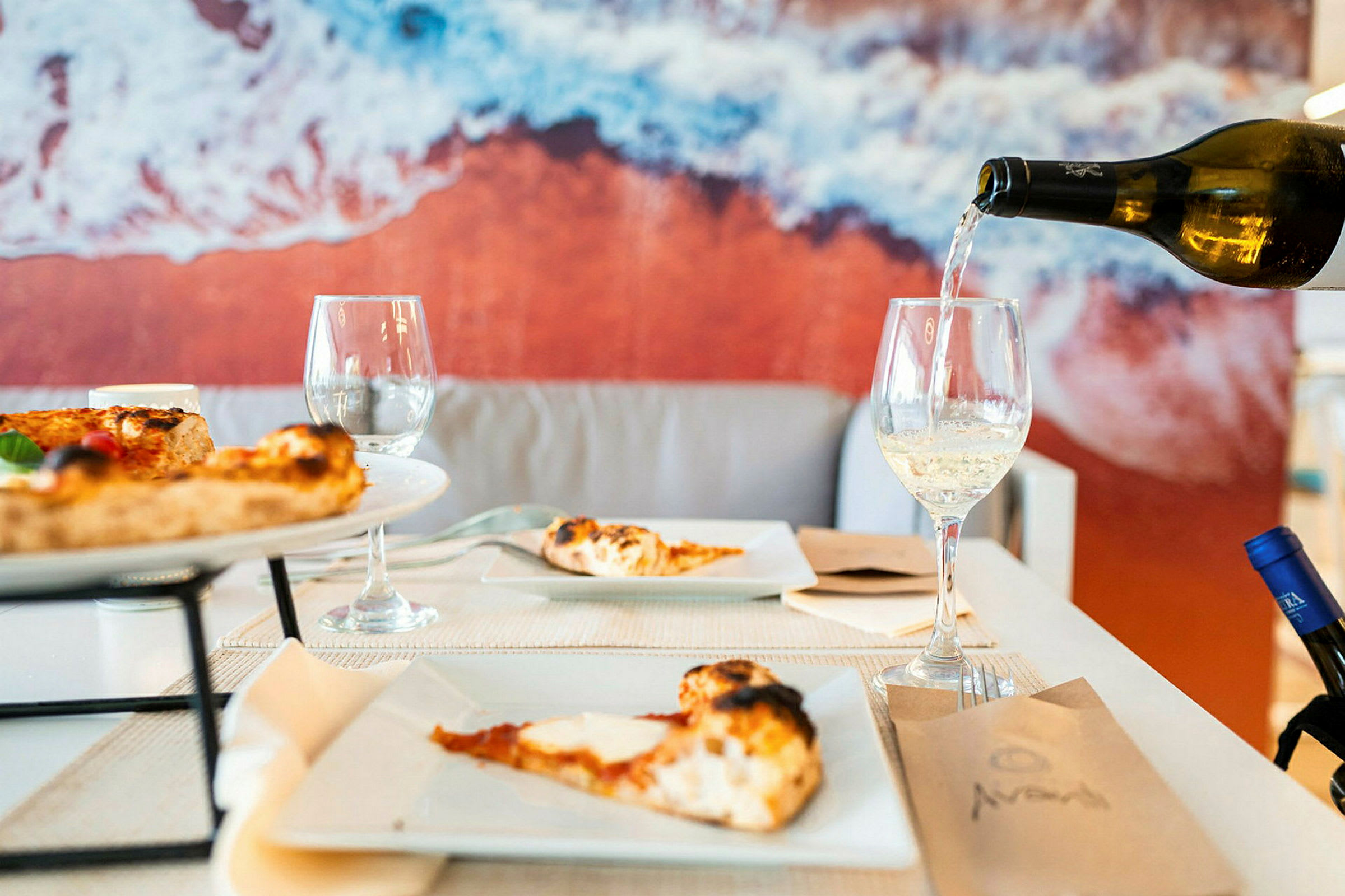Et bord med pizza og vinglas, baggrund med et billede af stranden. (AI-genereret tekst)