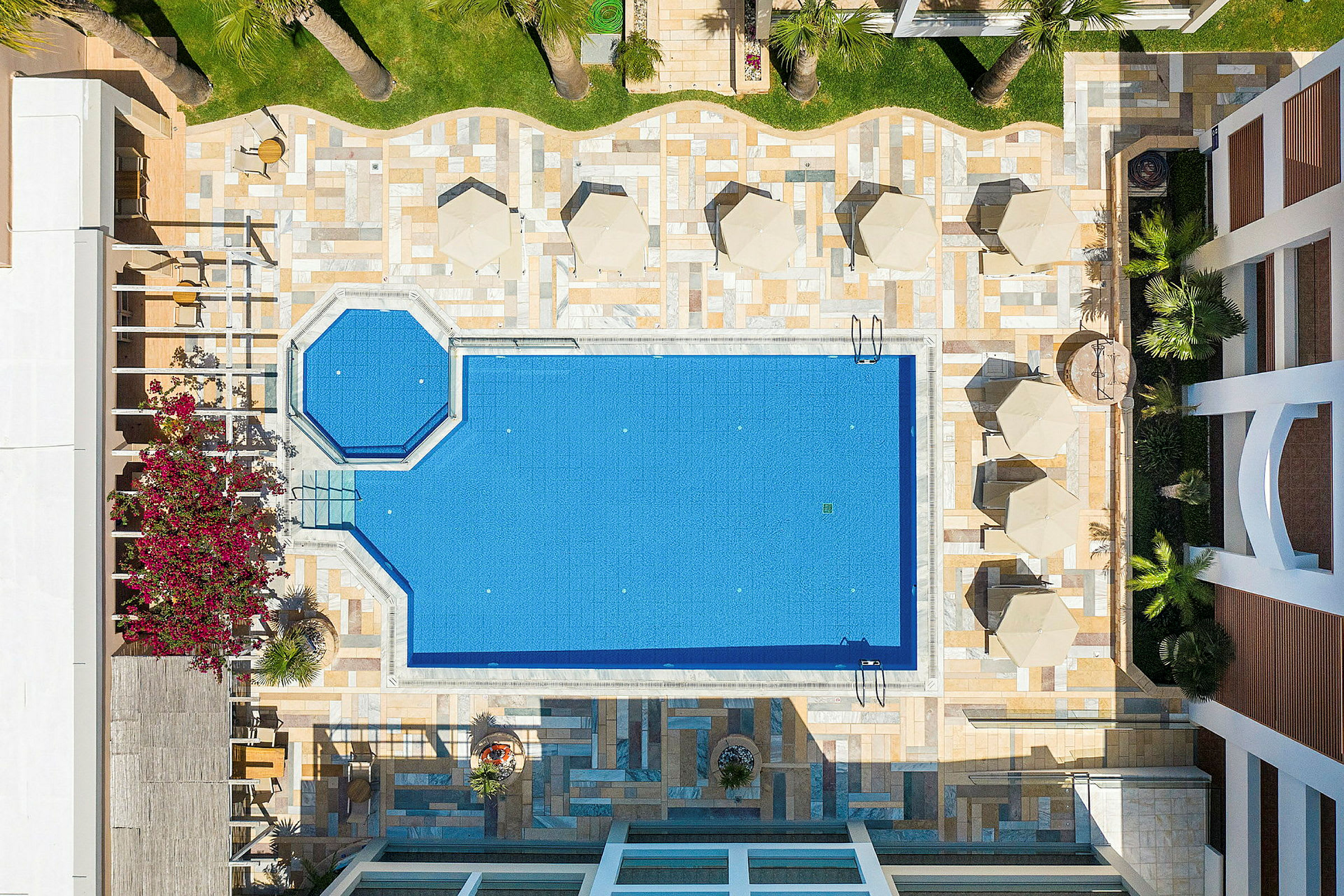 Billedet viser en udendørs swimmingpool omgivet af liggestole og parasoller, fotograferet fra oven. (AI-genereret tekst)