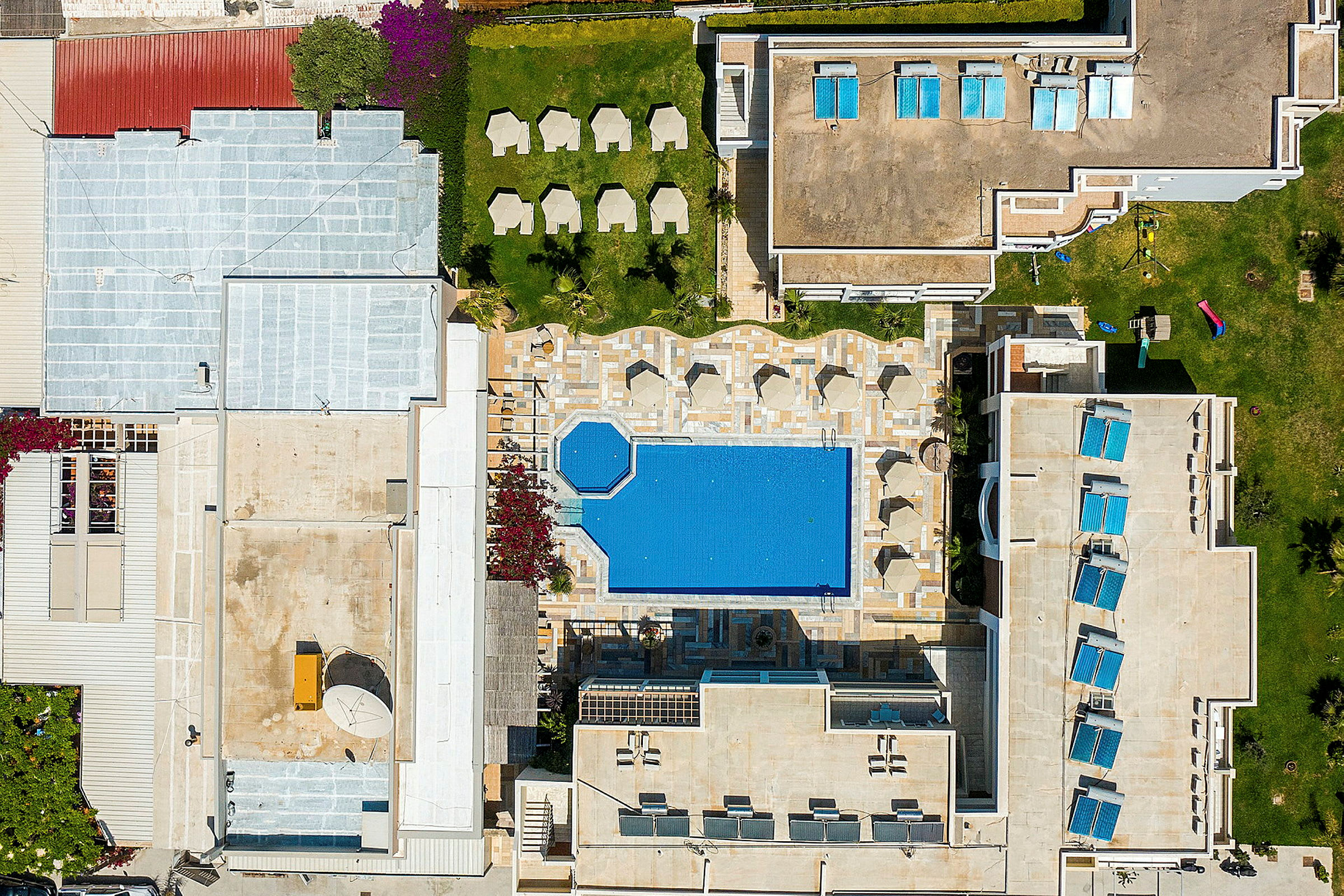 Luftfoto af en hotelpool omgivet af liggestole og bygninger. (AI-genereret tekst)