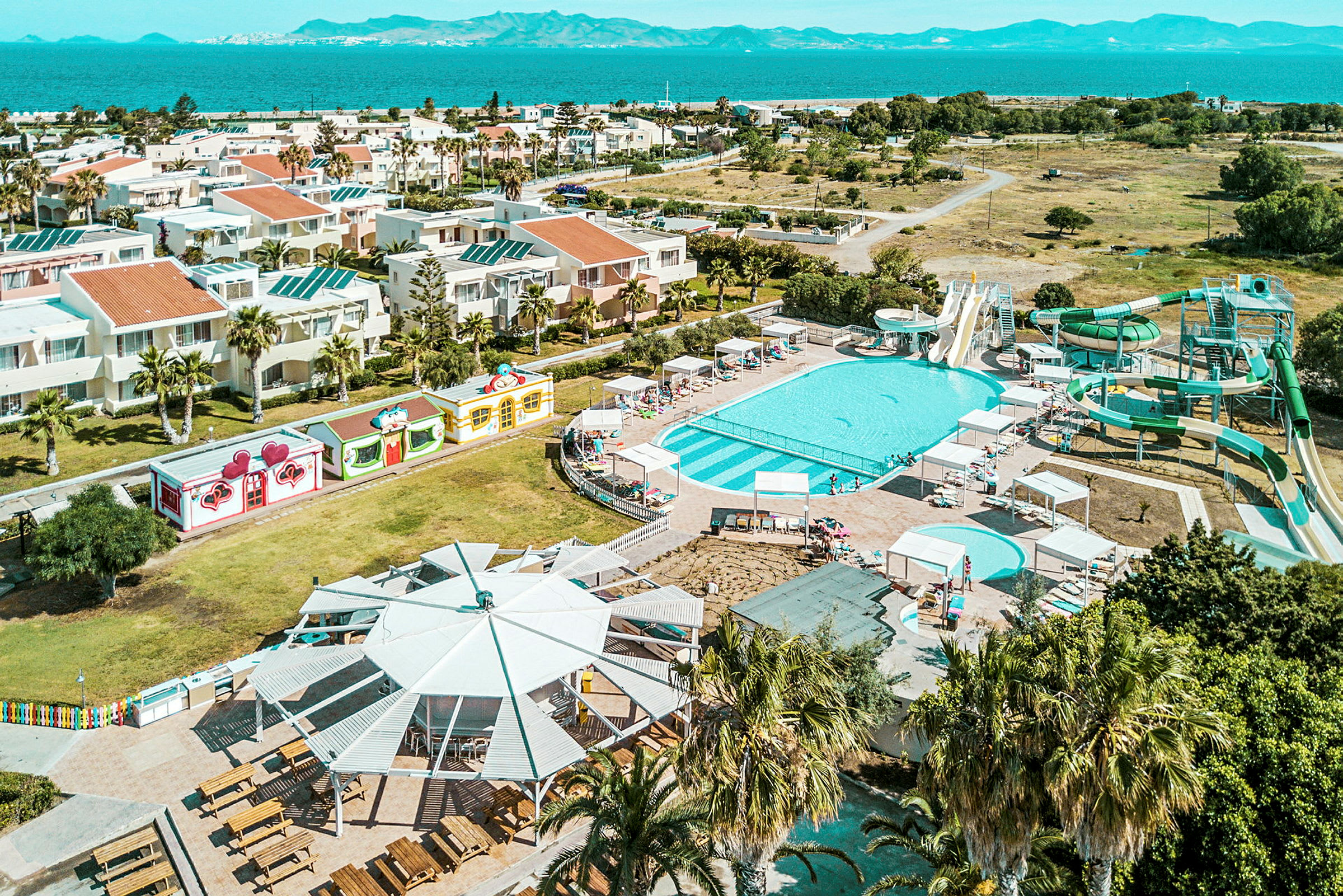 Luftfoto af et hotelområde med pools og vandrutsjebaner, omgivet af bygninger og grønne områder nær havet. (AI-genereret tekst)