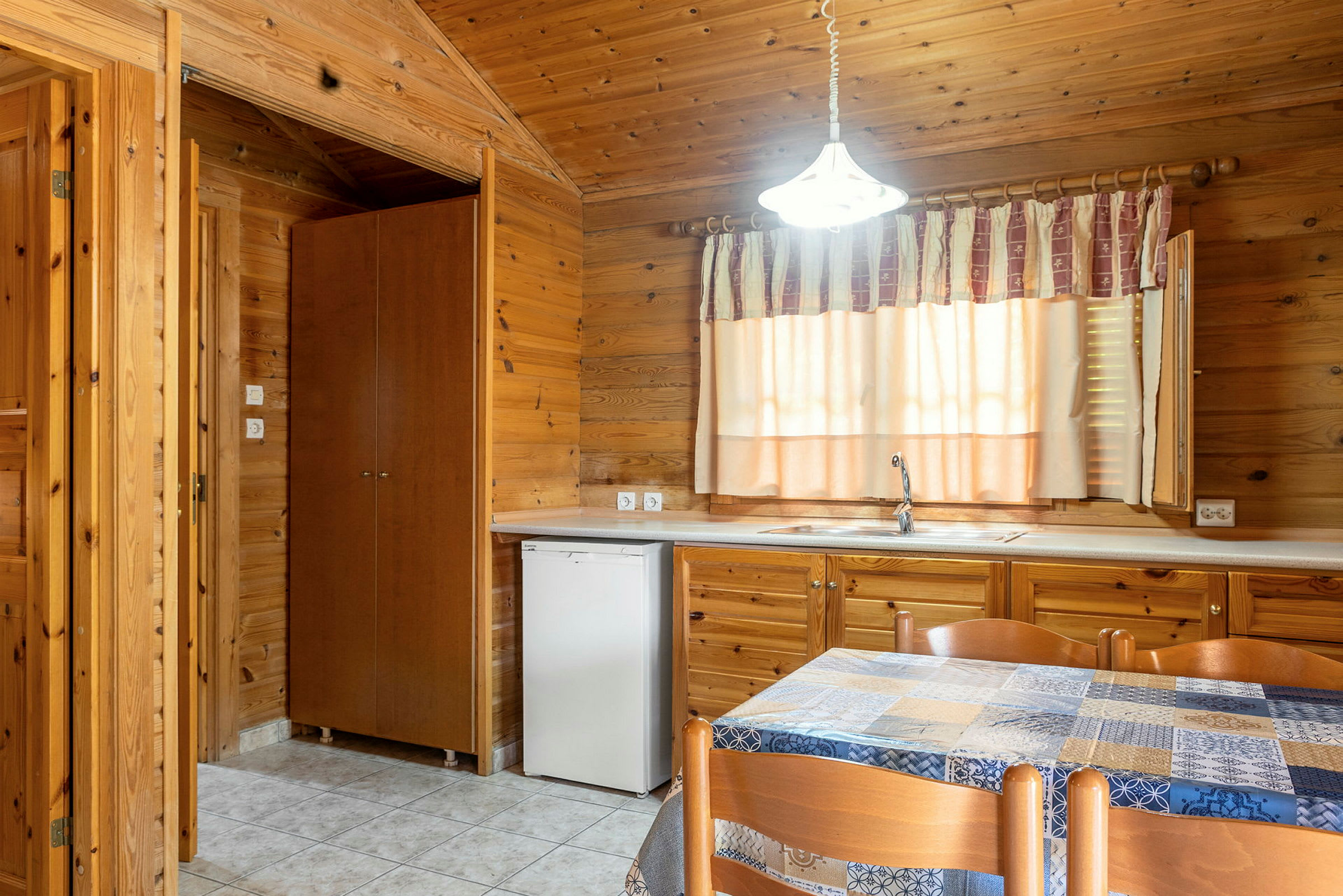 Indendørsmiljø med træpaneler, en køkkenvask med vandhane, et vindue med gardin og et bord med stole. (AI-genereret tekst)