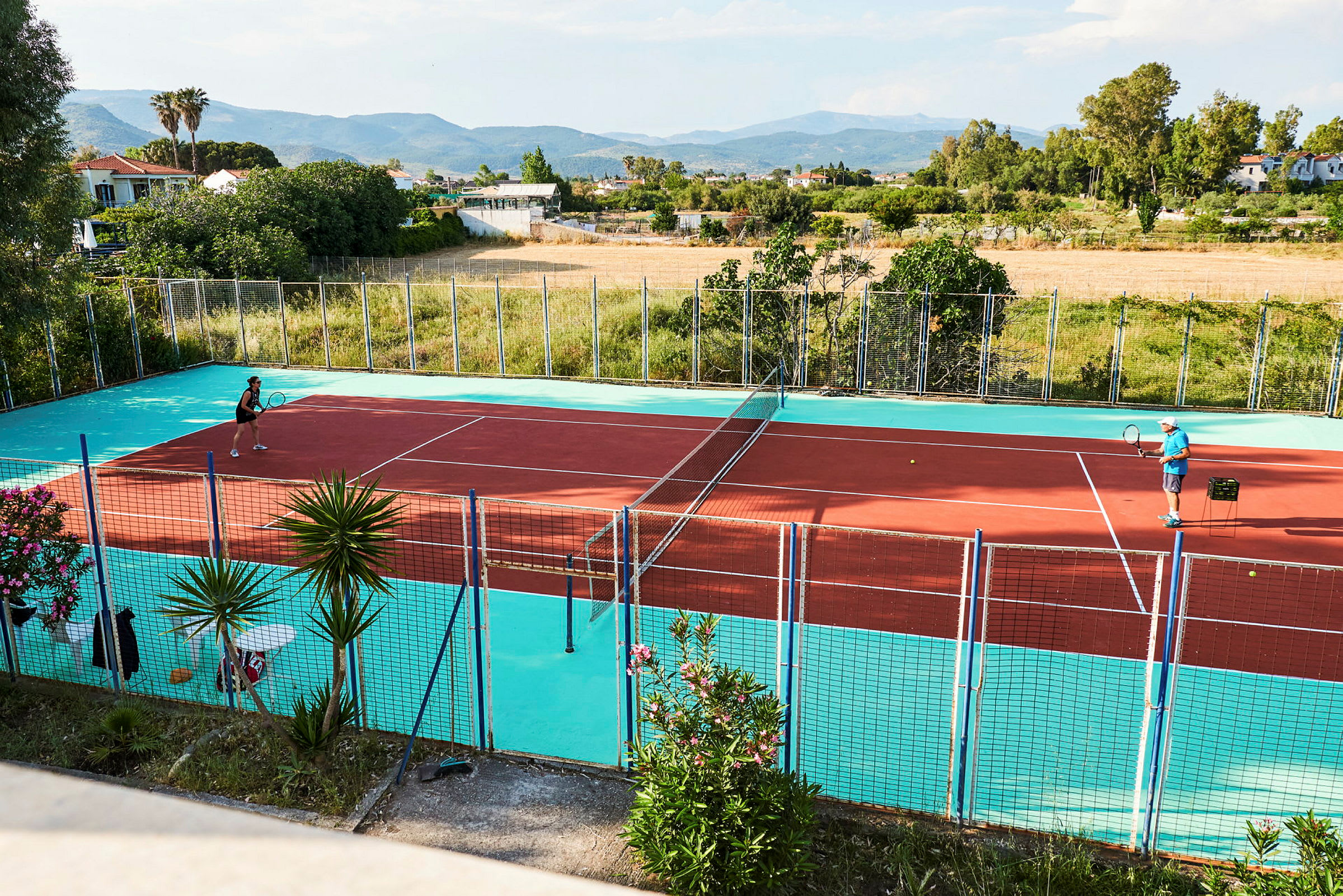To personer spiller tennis på en farverig tennisbane udendørs med landskab i baggrunden. (AI-genereret tekst)