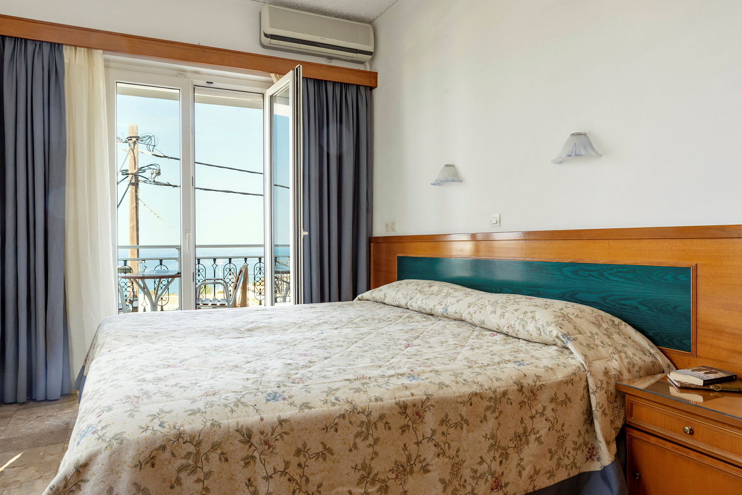Hotelværelse med en dobbeltseng og åbent vindue med udsigt til havet. (AI-genereret tekst)
