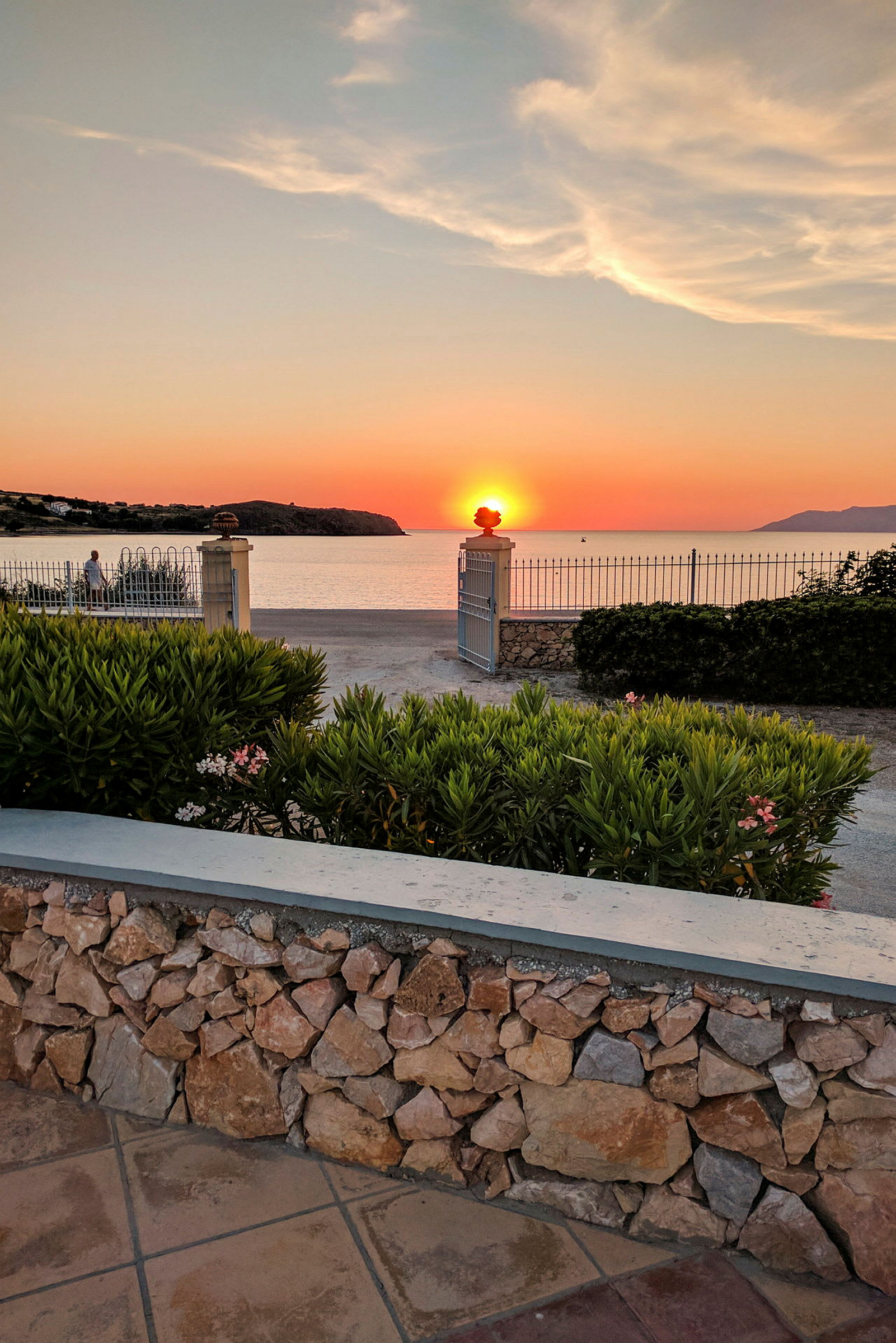 Solnedgang set over havet med en stenmursramme og vegetation i forgrunden. (AI-genereret tekst)