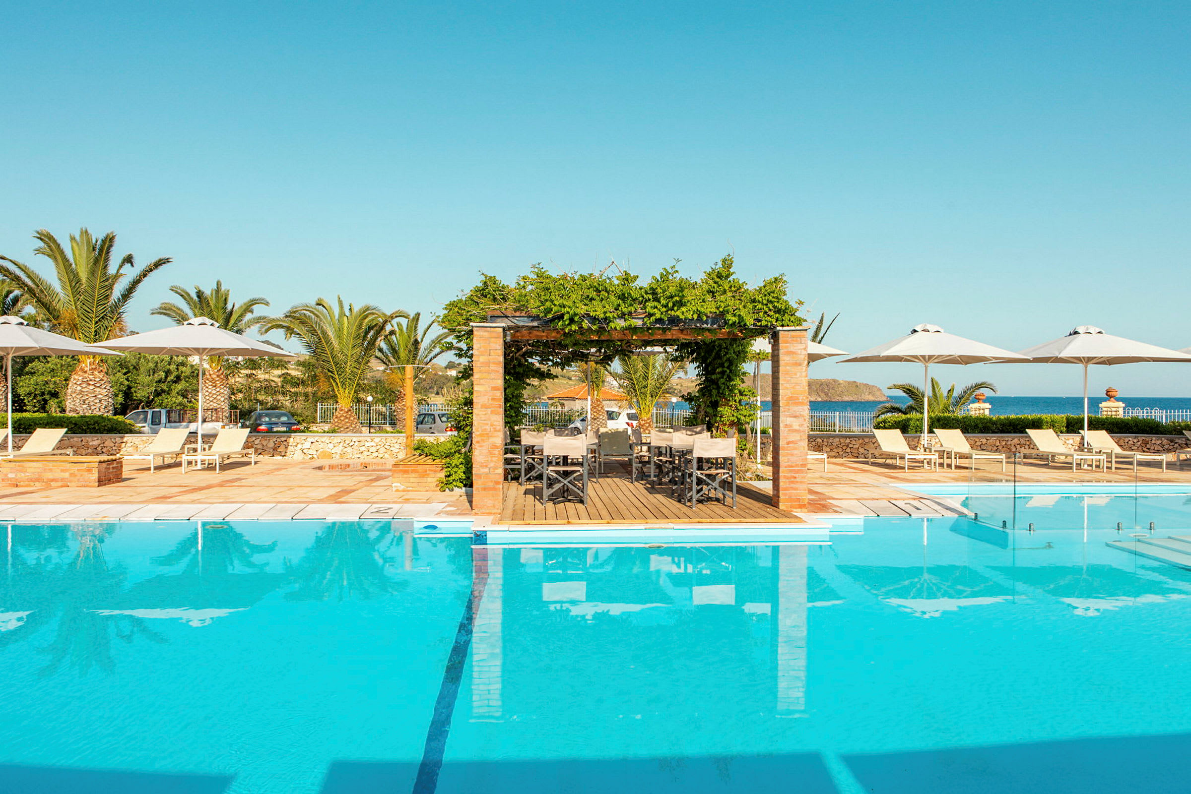 En swimmingpool omgivet af liggestole, parasoller og en pergola ved kysten. (AI-genereret tekst)