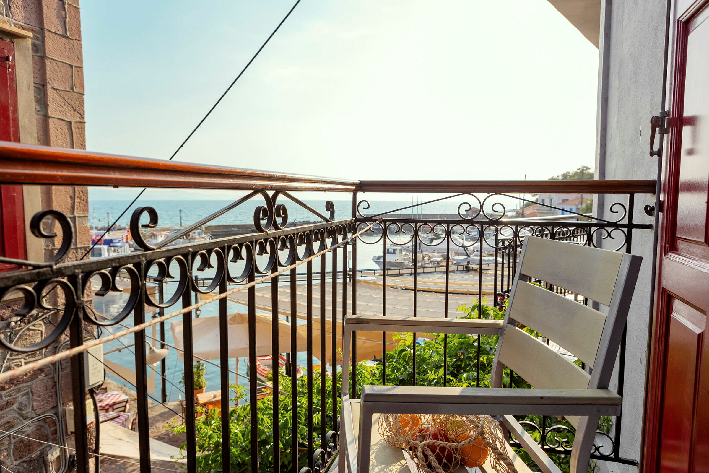Udsigt fra en balkon med smedejernsgelænder over en havn ved havet, en stol og et bord med net og frugt er på balkonen. (AI-genereret tekst)