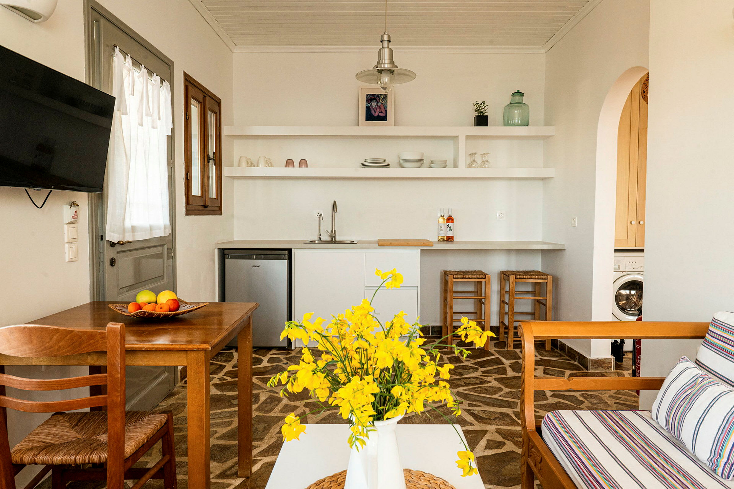 Et lyst indrettet køkken med hvide hylder, et træbord, gule blomster i en hvid vase og en stue med en træsofa og stribede puder. (AI-genereret tekst)