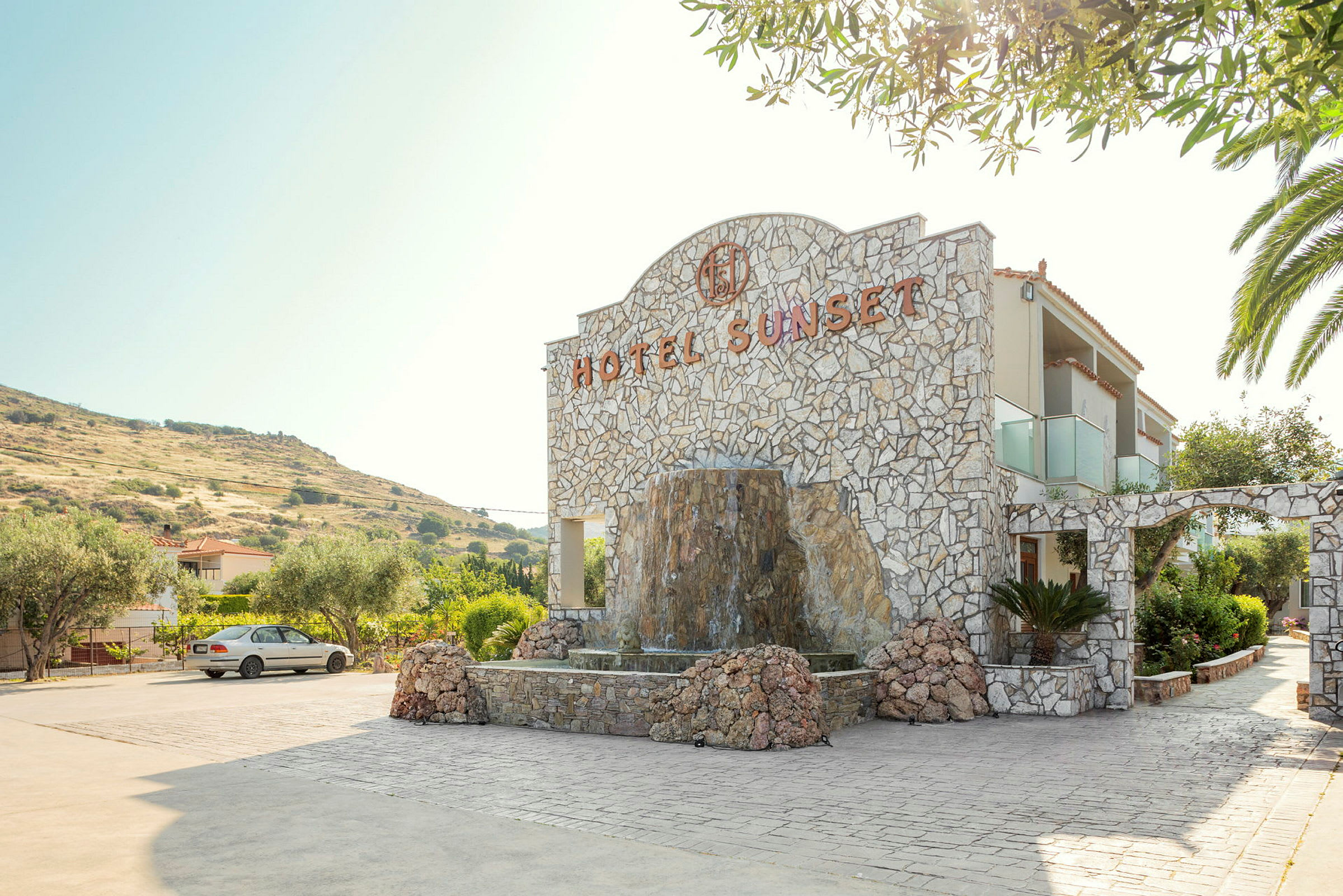 En stenbygning markeret med 'Hotel Sunset' og omgivet af en fontæne og grønt område. (AI-genereret tekst)