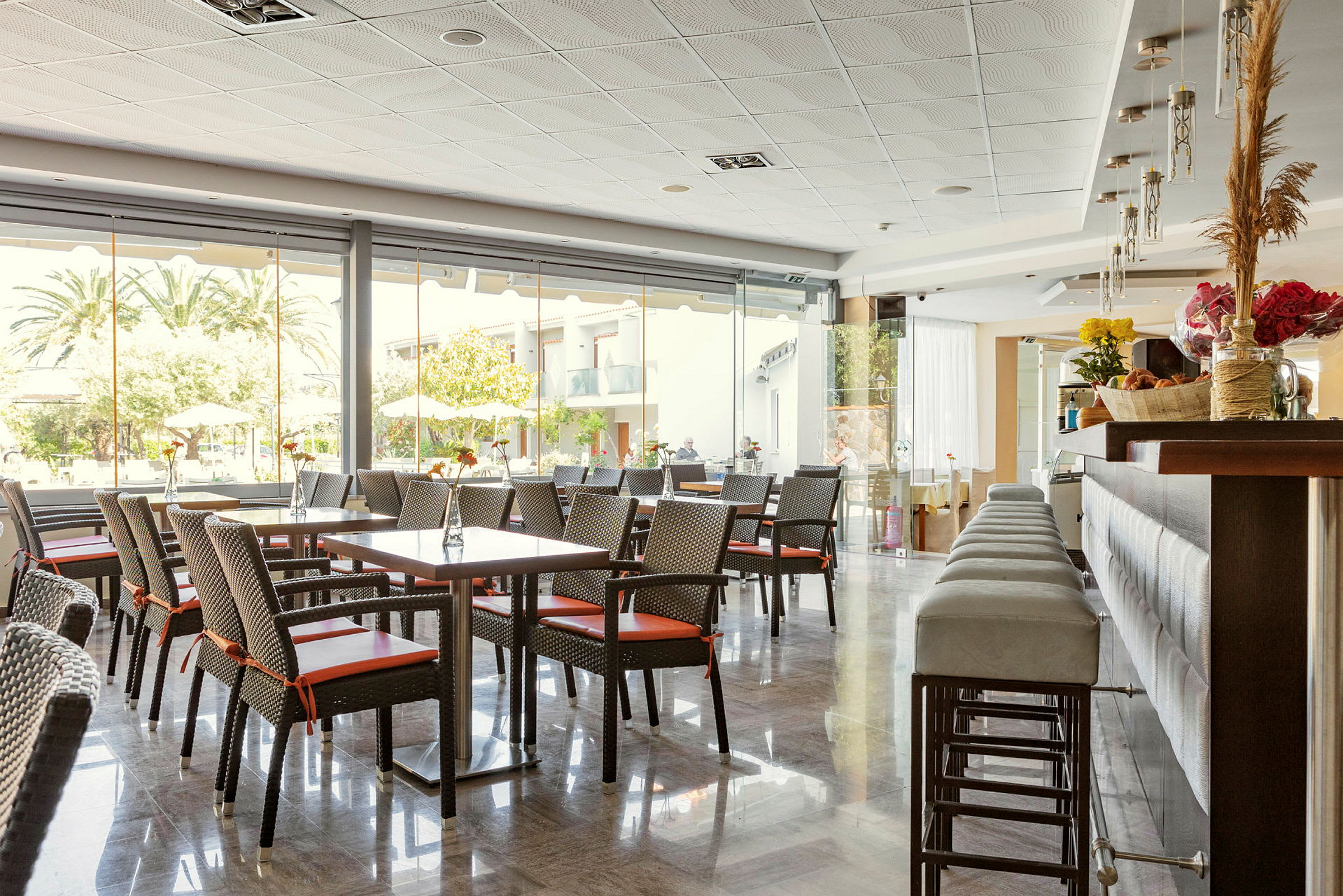 En hotelrestaurant med borde og stole samt en bardisk, store vinduer mod udendørs. (AI-genereret tekst)