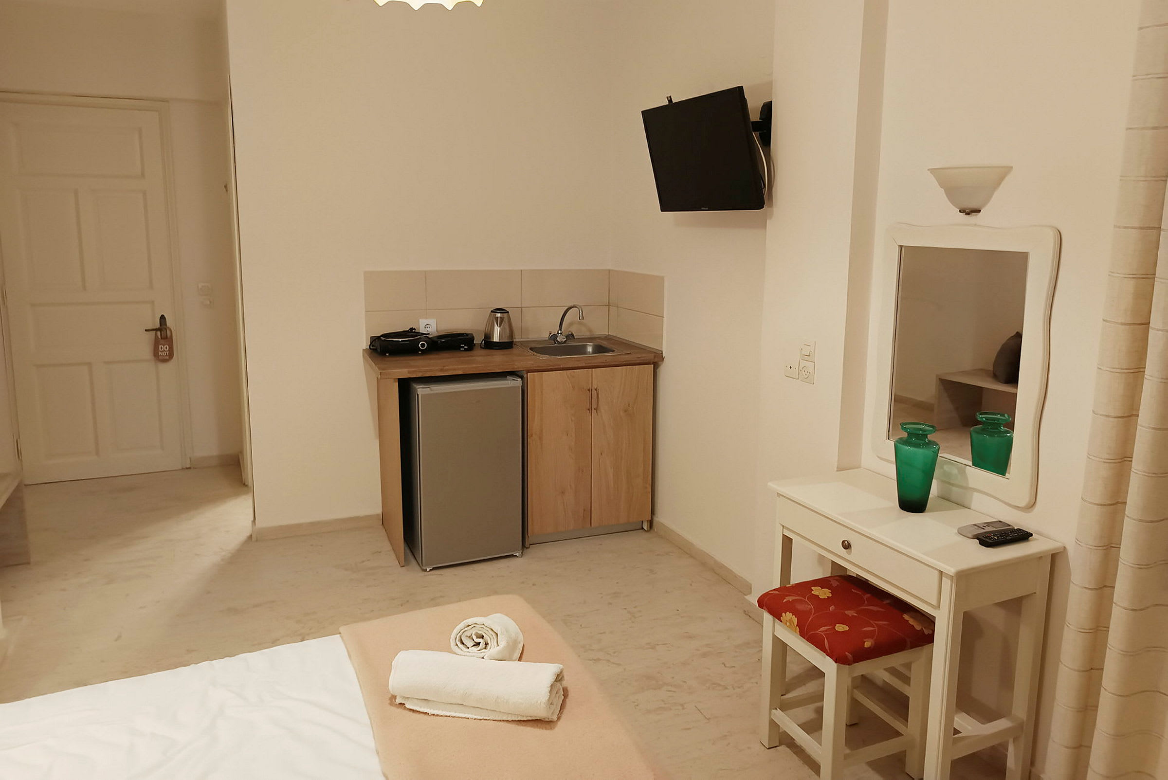 Hotelværelse med en lille køkkenafdeling, et køleskab, en vask og en komfur. Et skrivebord med spejl og stol samt en redt seng. (AI-genereret tekst)