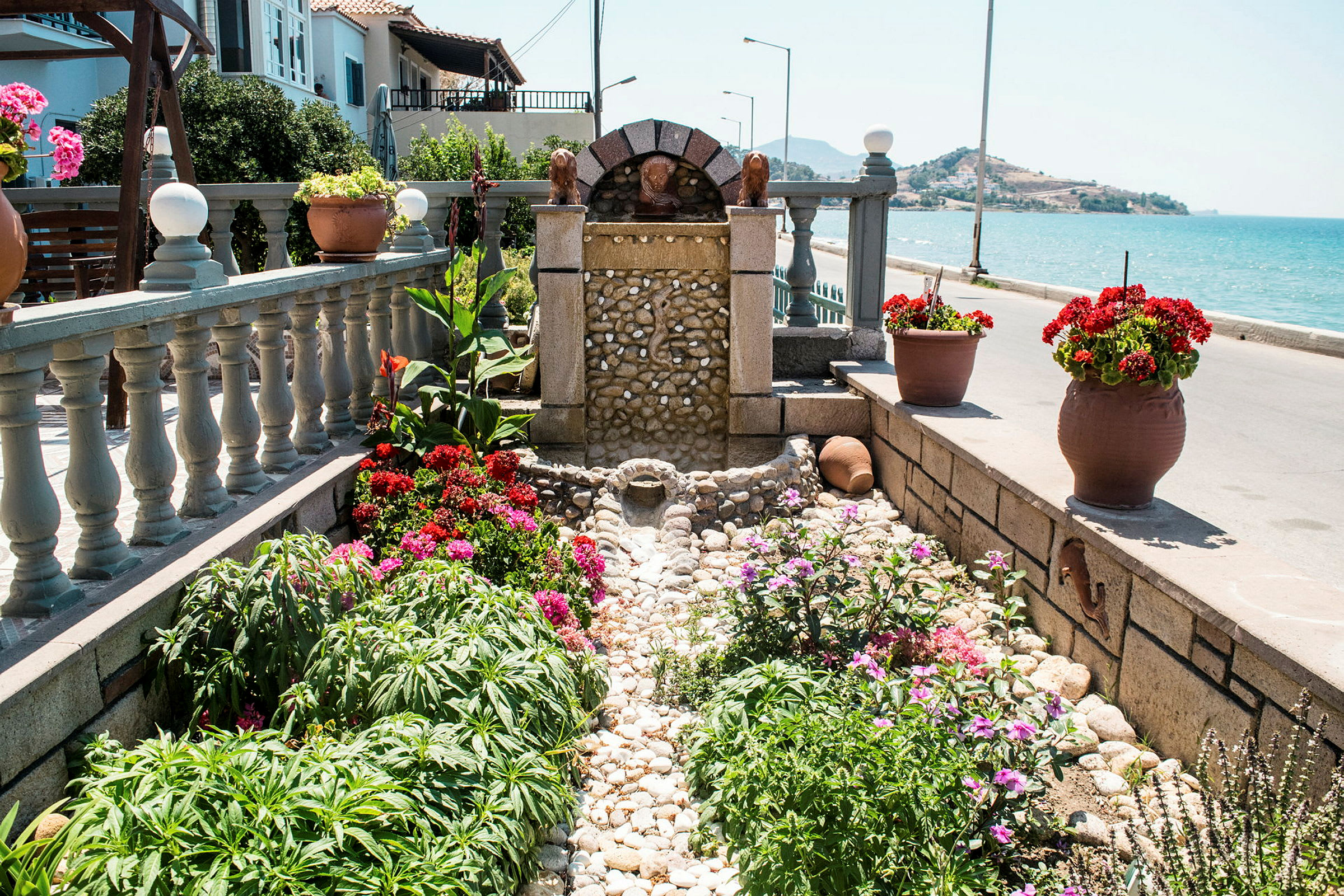 En lille fontæne omgivet af blomster og planter ved havet. (AI-genereret tekst)