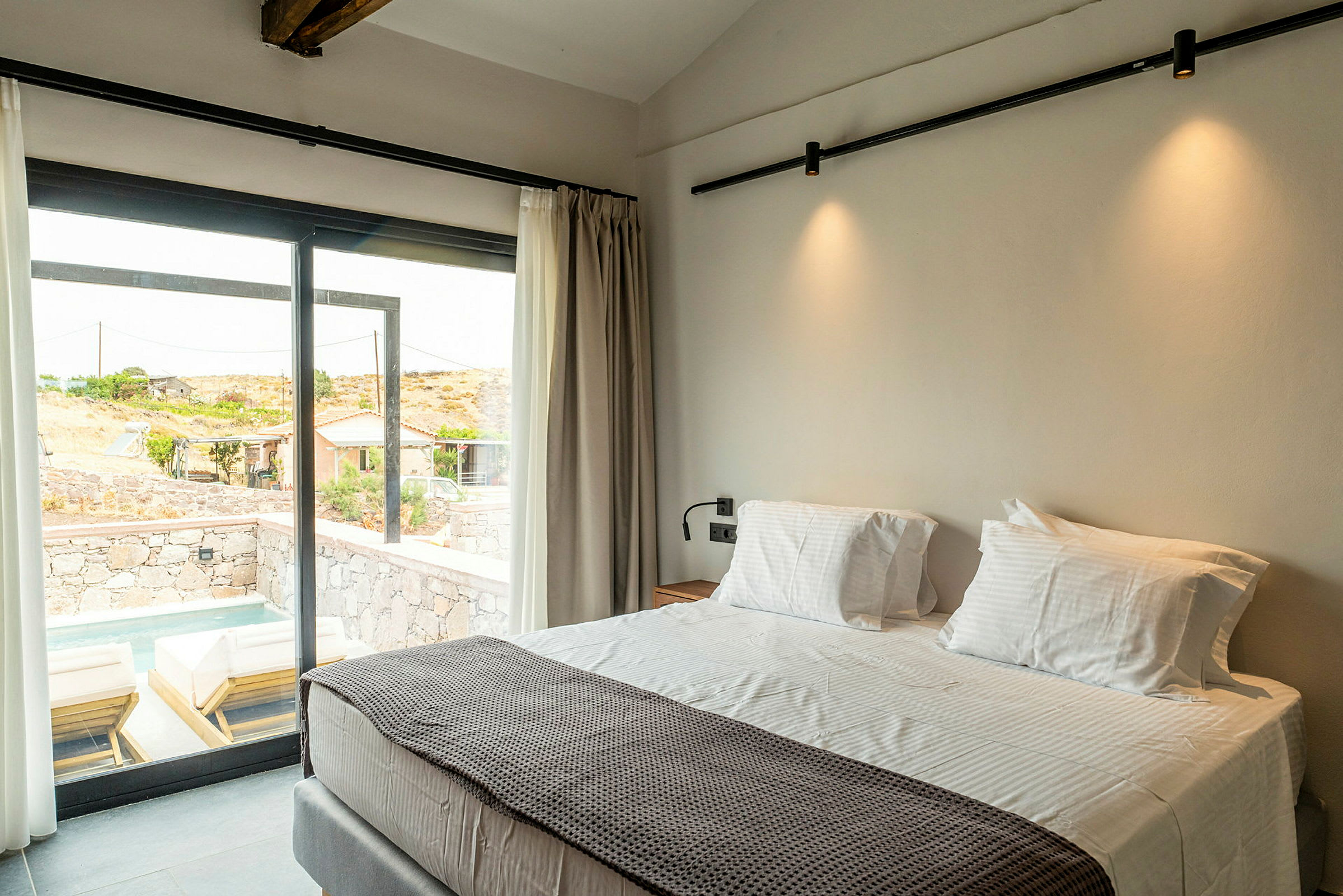 Hotelværelse med stor seng ved et vindue med udsigt til en terrasse med liggestole og en lille pool. (AI-genereret tekst)