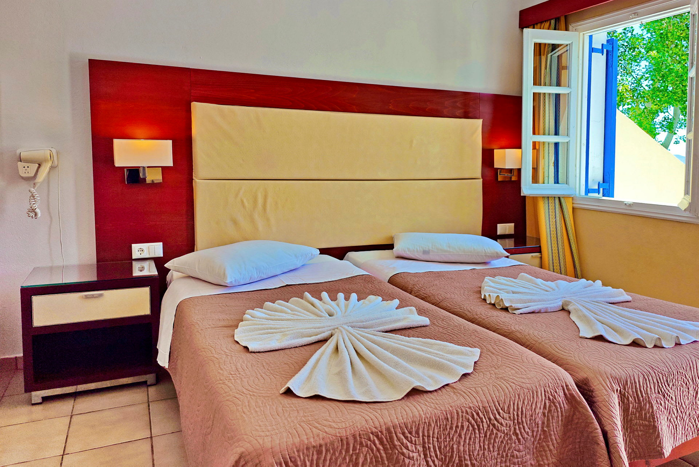 Hotelværelse med to enkeltsenge placeret side om side, redt med mønstrede sengetæpper og håndklæder foldet som dekorative figurer. Et natbord med telefon står ved siden af sengen, og et åbent vindue lukker dagslyset ind. (AI-genereret tekst)