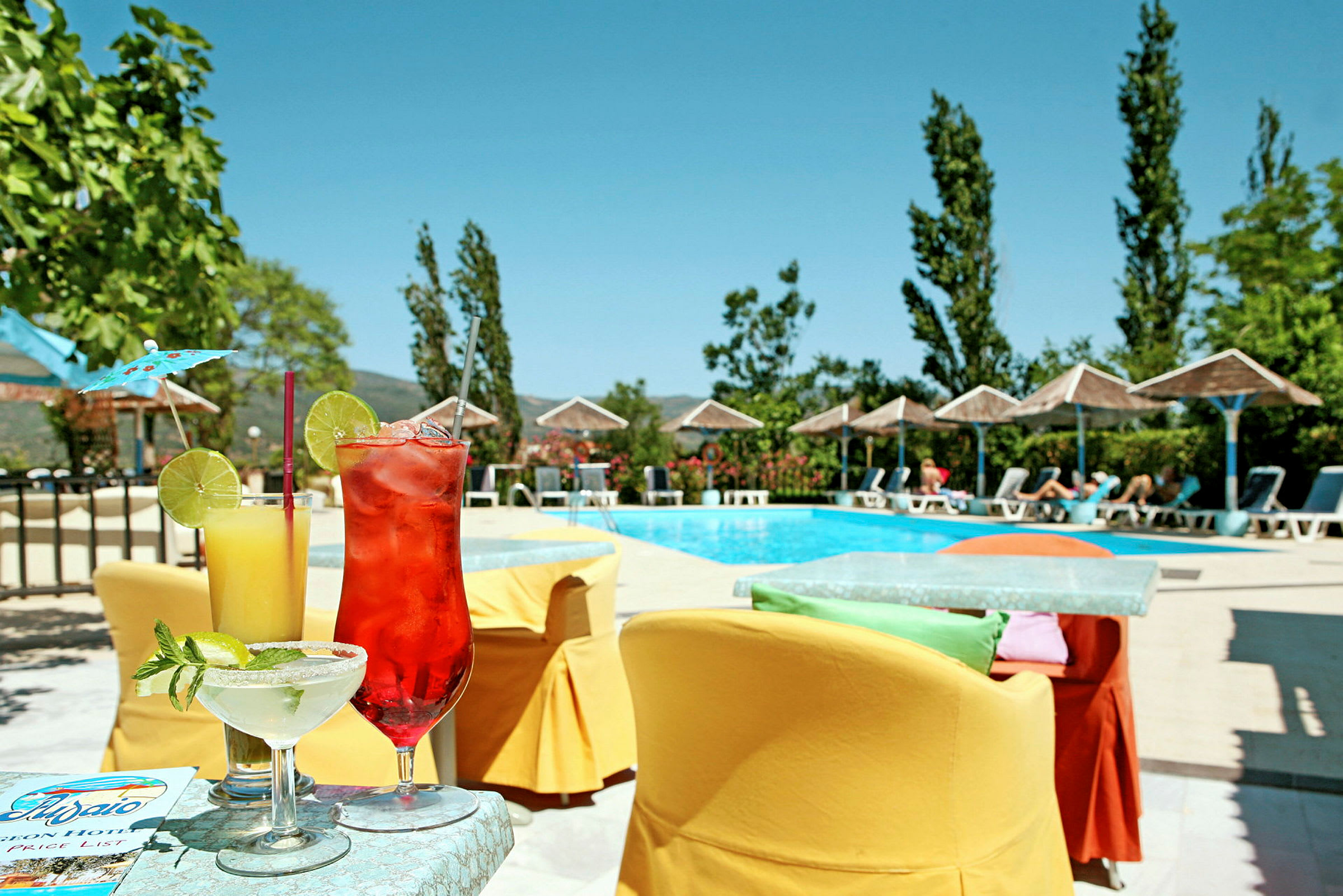 Billede af en pool omgivet af liggestole og parasoller. På et bord i forgrunden står tre farverige drinks. (AI-genereret tekst)