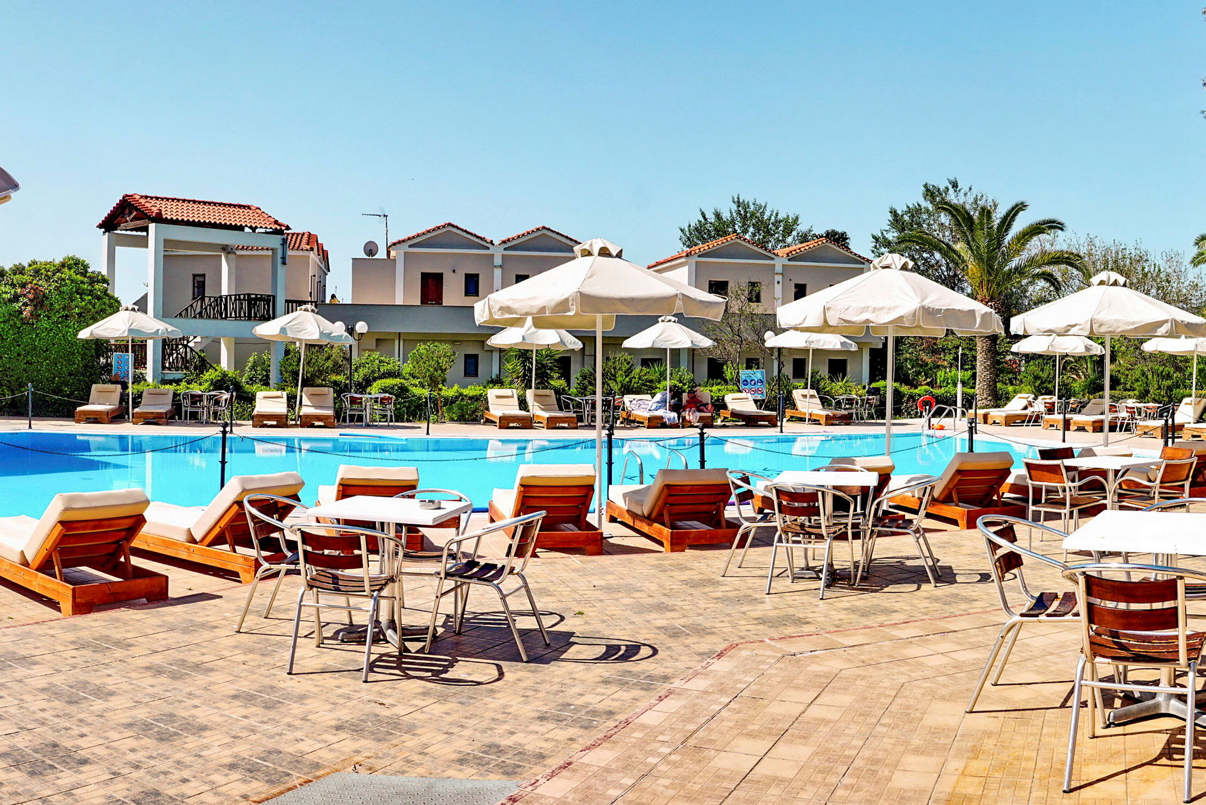 En hotelpool omgivet af liggestole, parasoller og bygninger. (AI-genereret tekst)