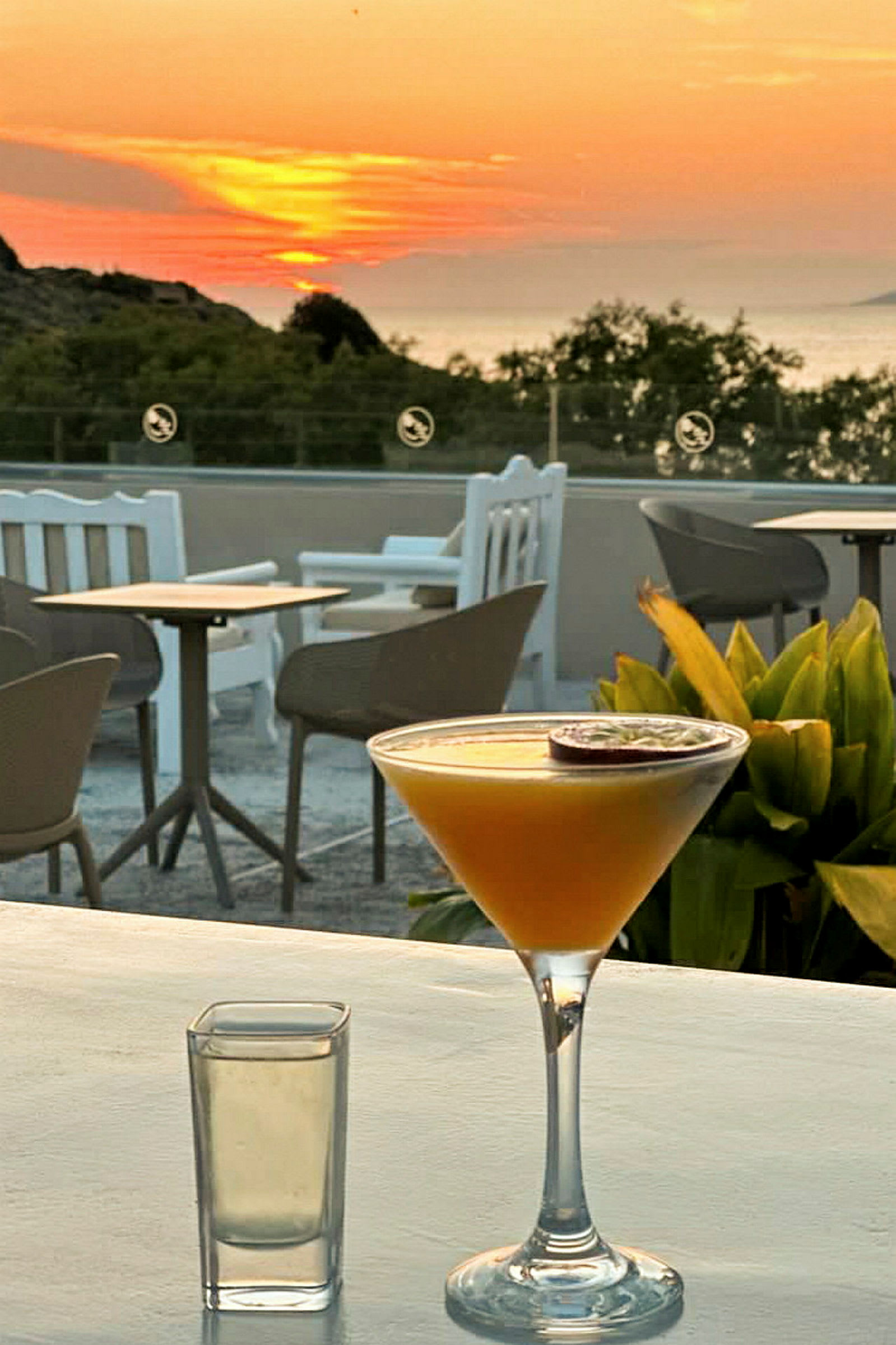 En tropisk cocktail og et glas står på et bord ved solnedgang med siddeområder og beplantning i baggrunden. (AI-genereret tekst)