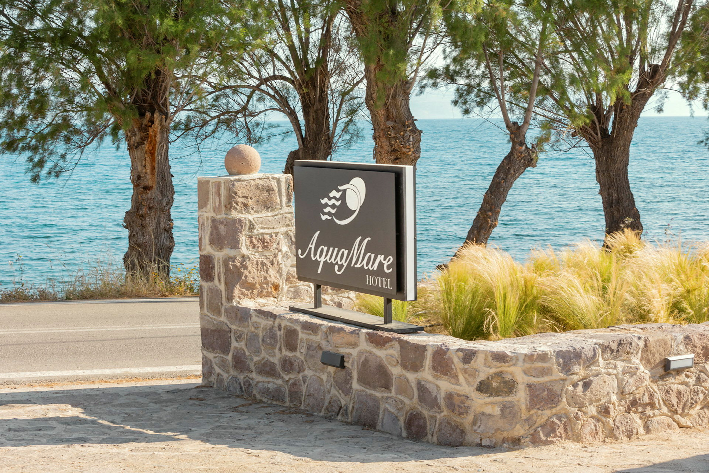 Hoteltskilt med teksten 'Aqua Mare Hotel', omgivet af stenmur og planter, med havet og træer i baggrunden. (AI-genereret tekst)