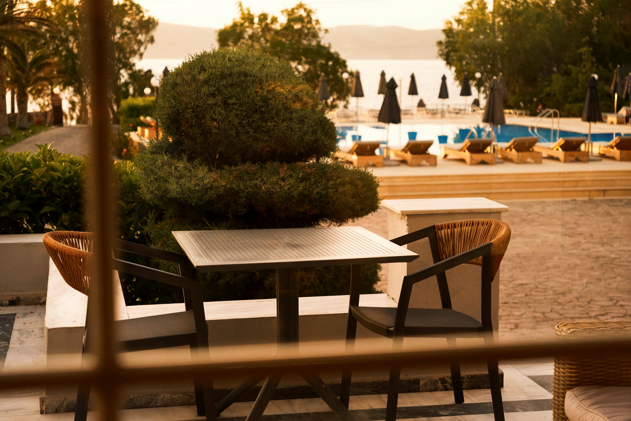 Et udendørs bord med to stole på en terrasse, med en pool og liggestole i baggrunden. (AI-genereret tekst)