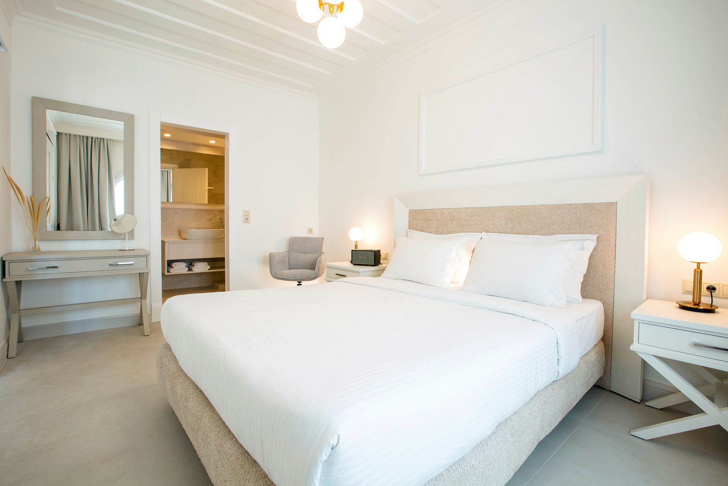 Et lyst og moderne hotelværelse med en stor seng, to natbord med lamper, et skrivebord med spejl og et åbent badeværelse. (AI-genereret tekst)
