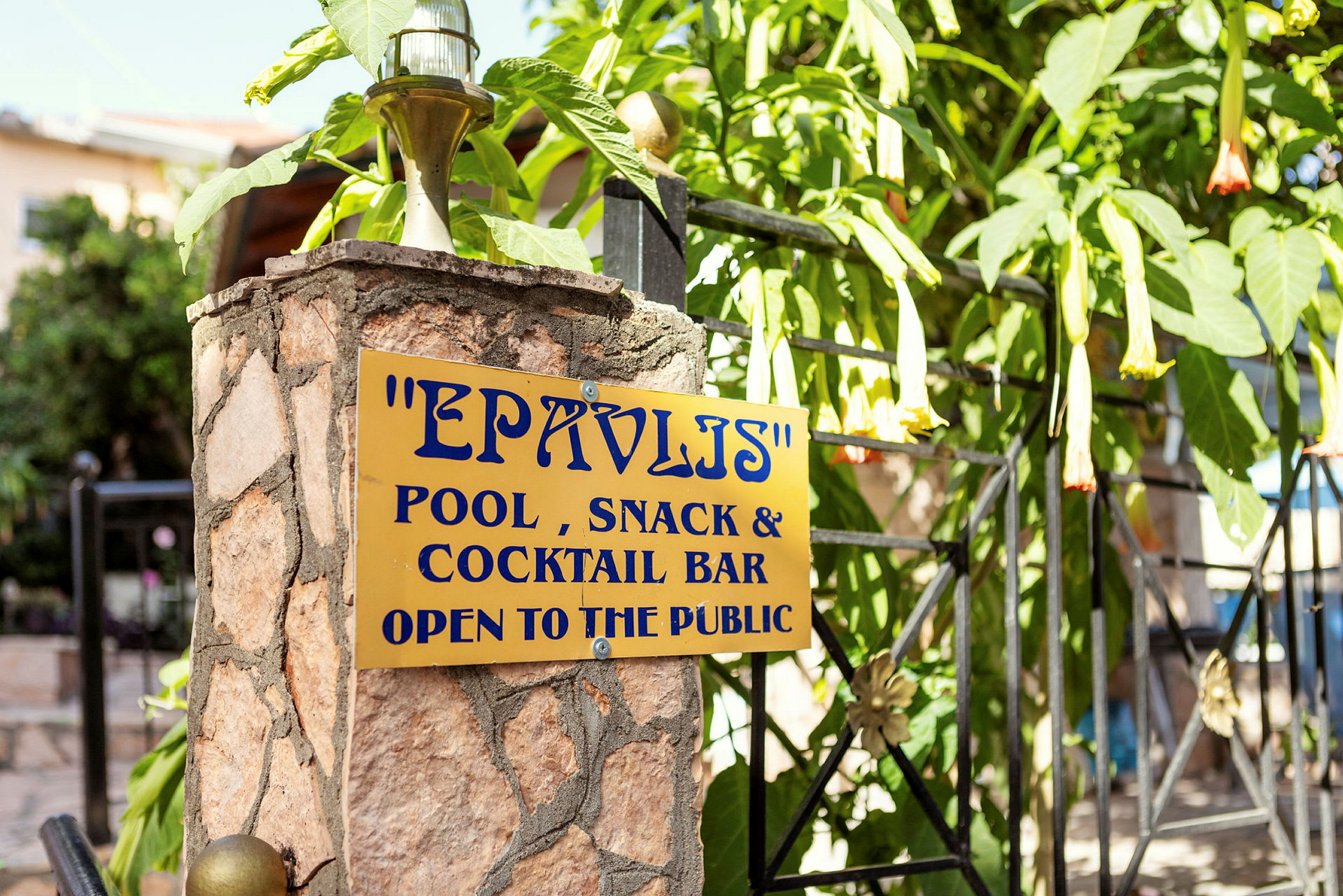 Skilt på en stensøjle med teksten 'EPAVLIS Pool, Snack & Cocktail Bar Open to the public'. (AI-genereret tekst)