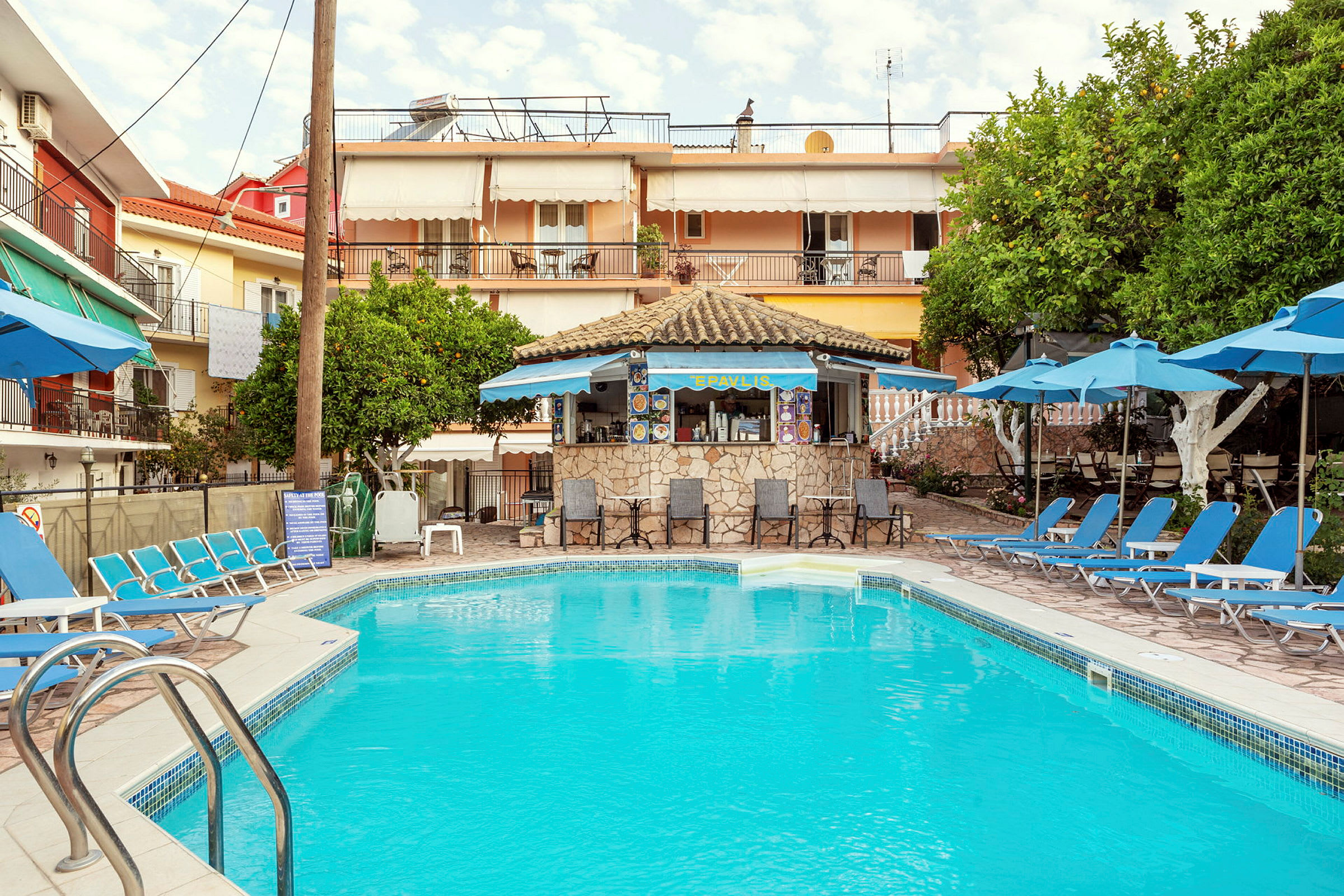 En udendørs swimmingpool omgivet af liggestole og parasoller, med en lille café i baggrunden. (AI-genereret tekst)