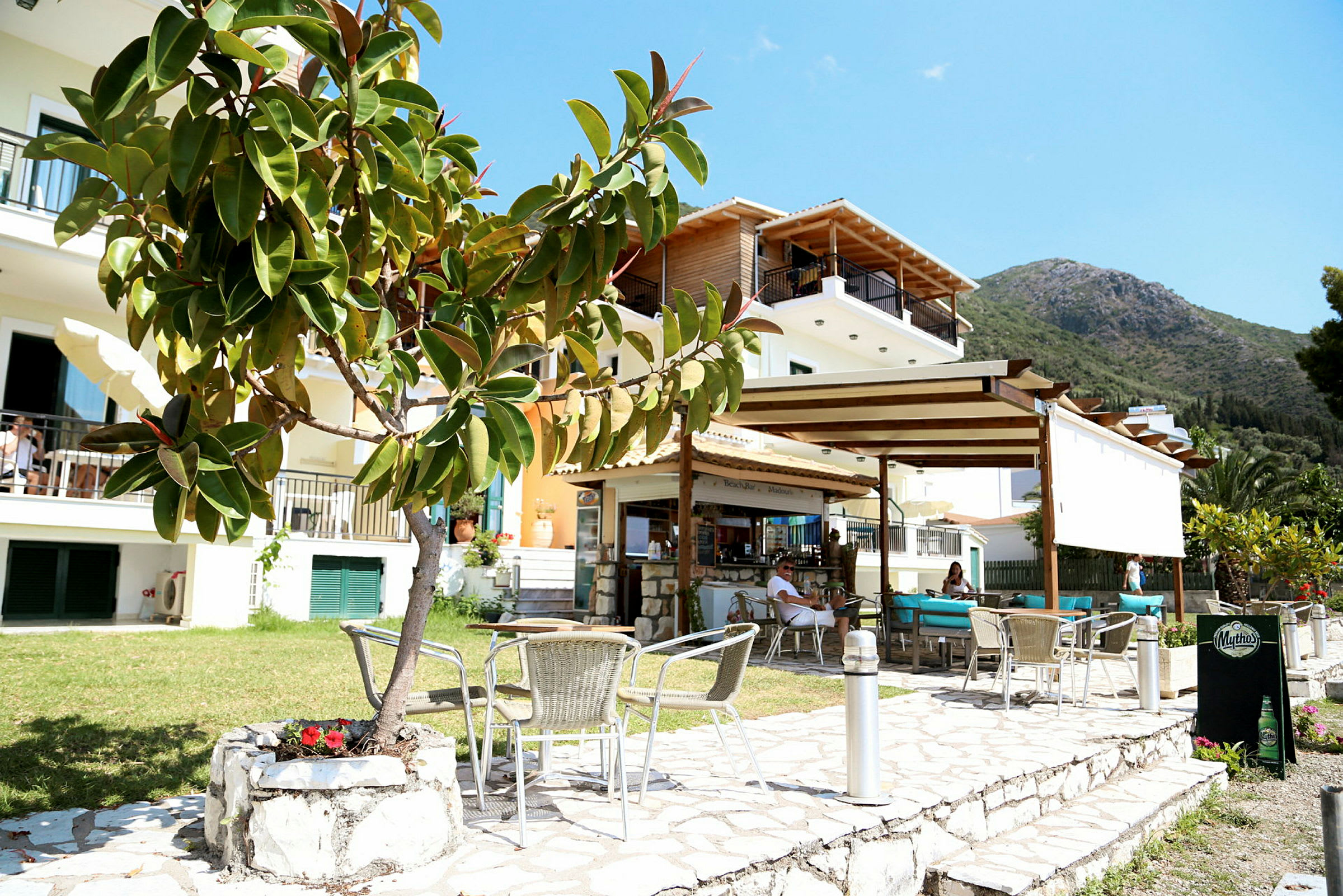 En udendørs restaurant foran et mindre lejlighedshotel med en bjergskråning i baggrunden. (AI-genereret tekst)